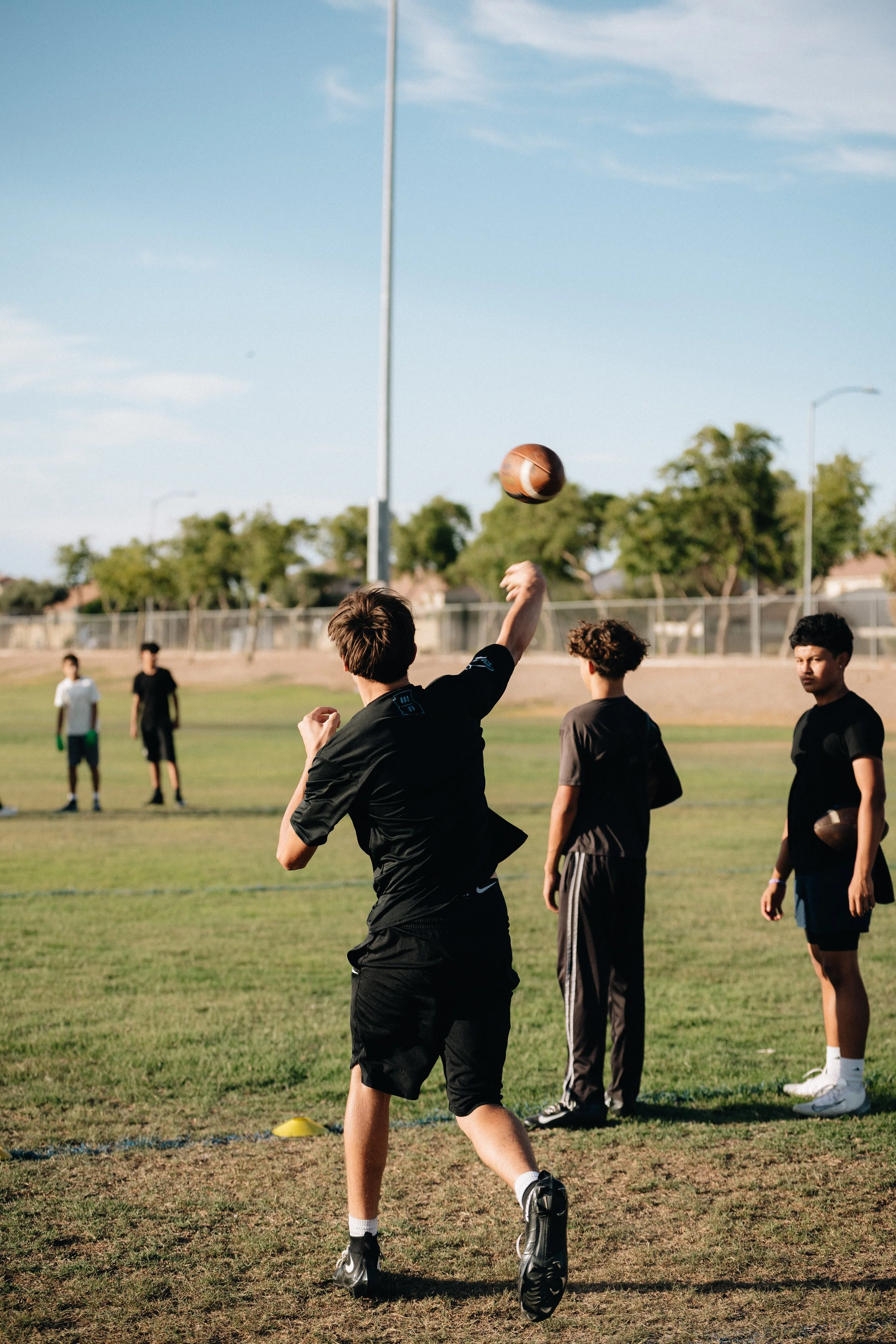 A group of young men playing football on a grassy field under a blue sky with few clouds. Some are standing, and one is in the process of catching or throwing a football. The scene appears to be during a sunny day.