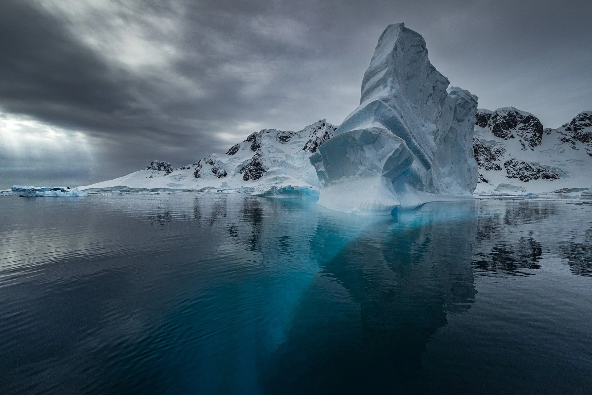 Iceberg floating in cold ocean water with snow-covered mountains and a cloudy sky in the background.