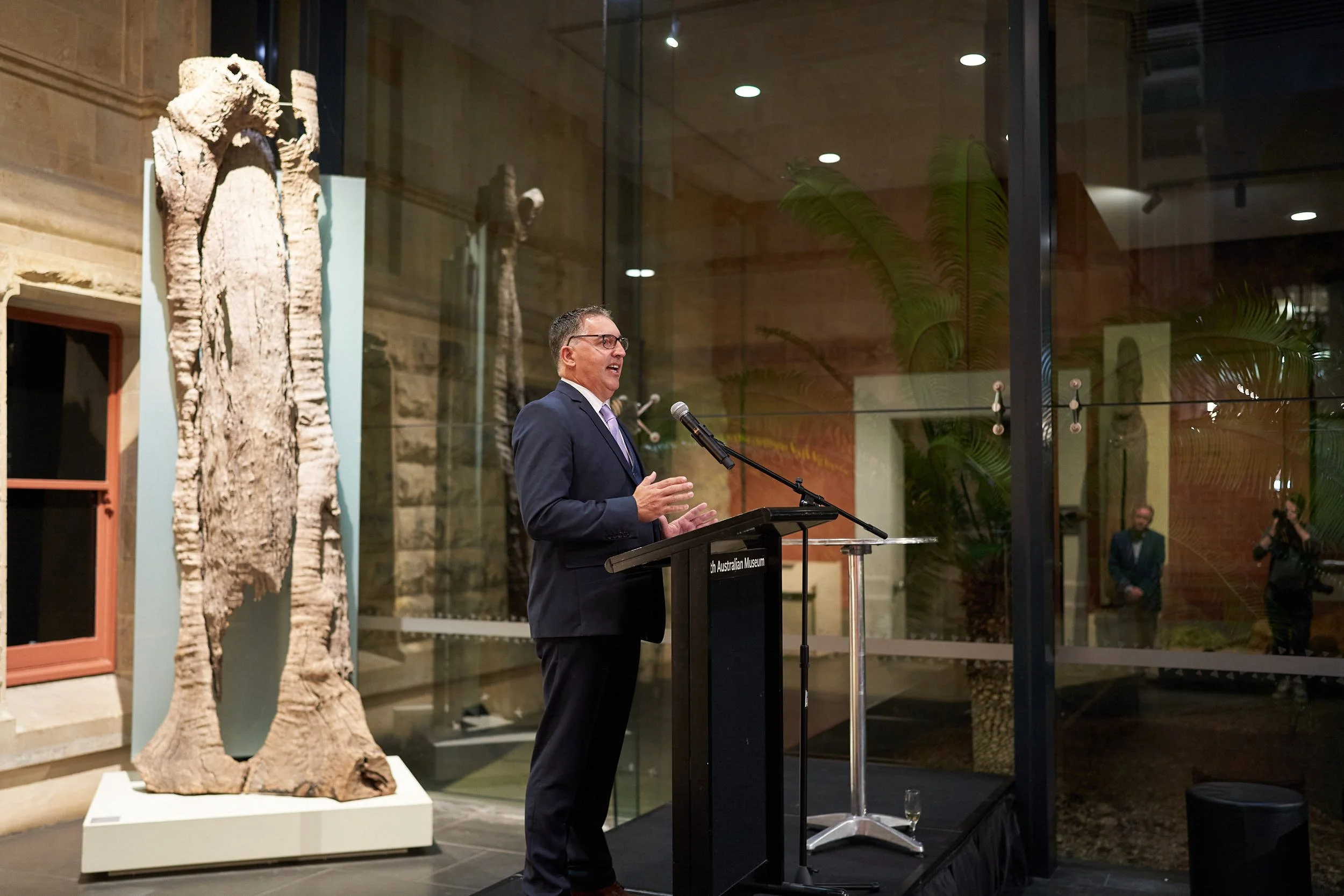 A man in a suit and glasses is giving a speech at a podium in an art museum, with fossil artifacts displayed behind him and glass walls showing trees outside.