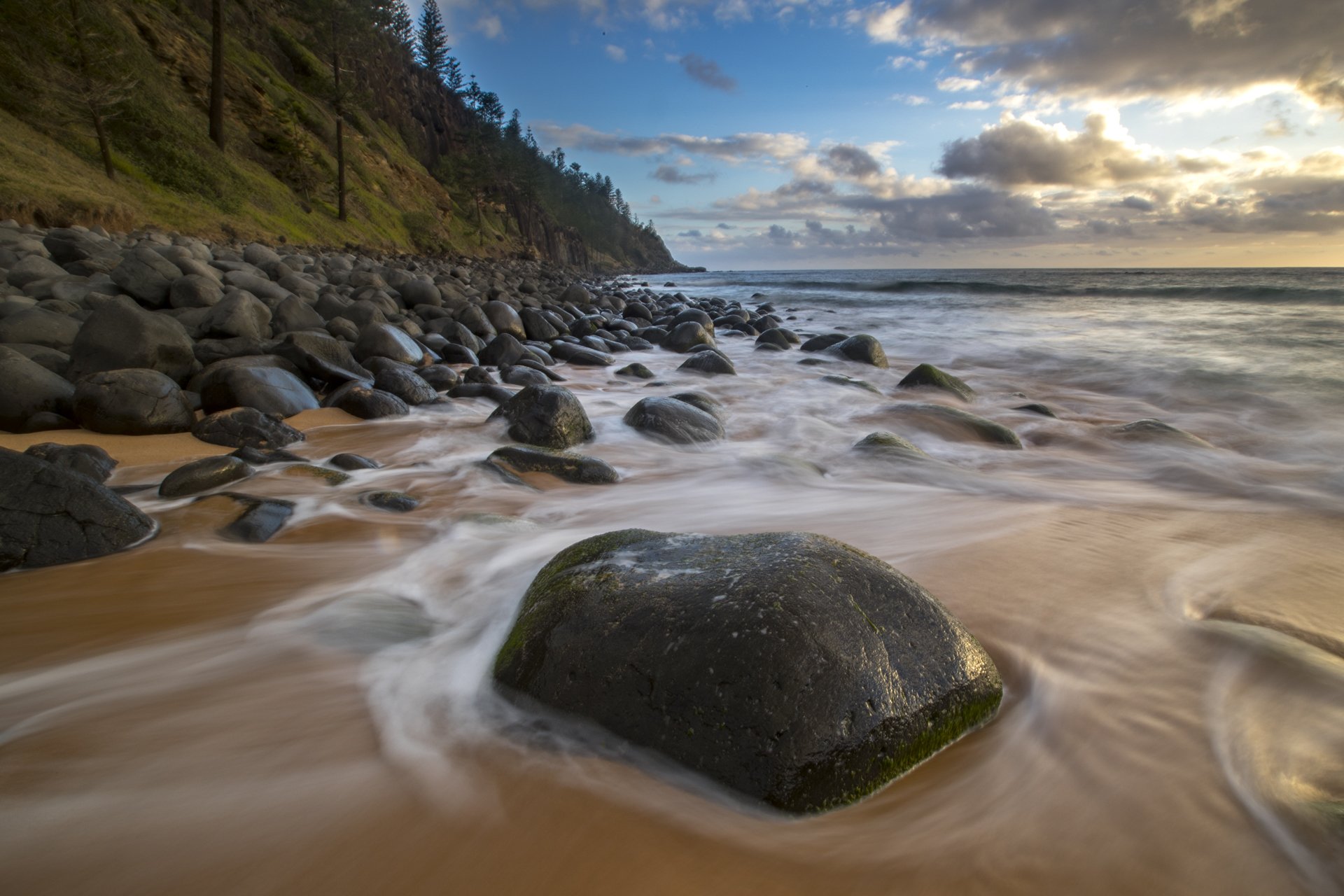 Rocky beach shoreline with waves washing over the sand and large moss-covered boulders, a forested hillside on the left, and a partly cloudy sky overhead.