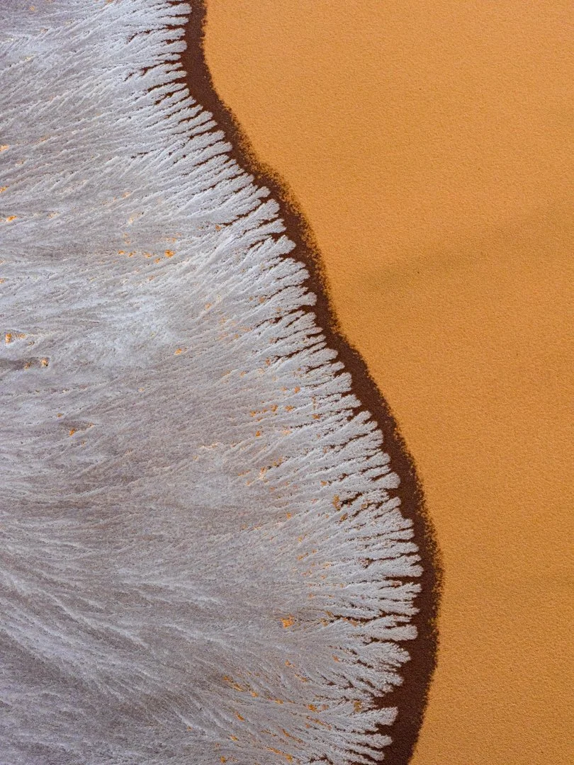 Close-up of an orange desert dune with snow or salt forming a white, feathered pattern along the edge.