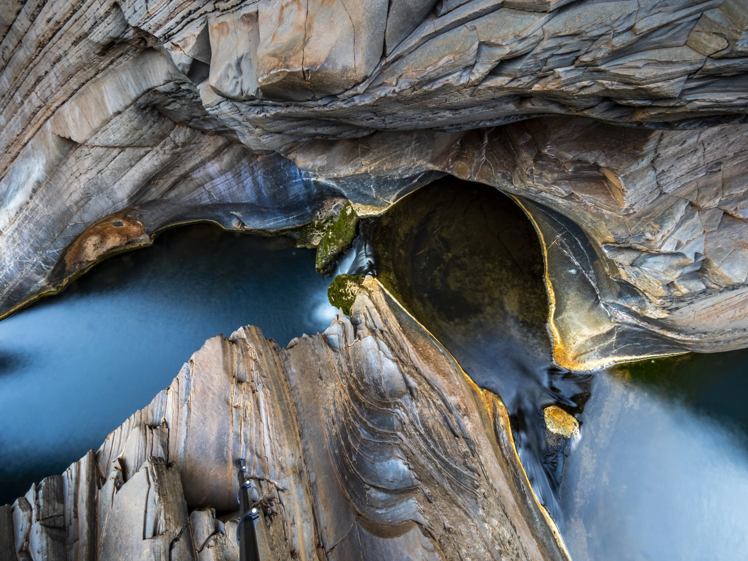 A deep canyon with layered rock walls and a river flowing through the bottom. The rocks have various shades of gray, brown, and orange, with some patches of green moss or plants near the water.