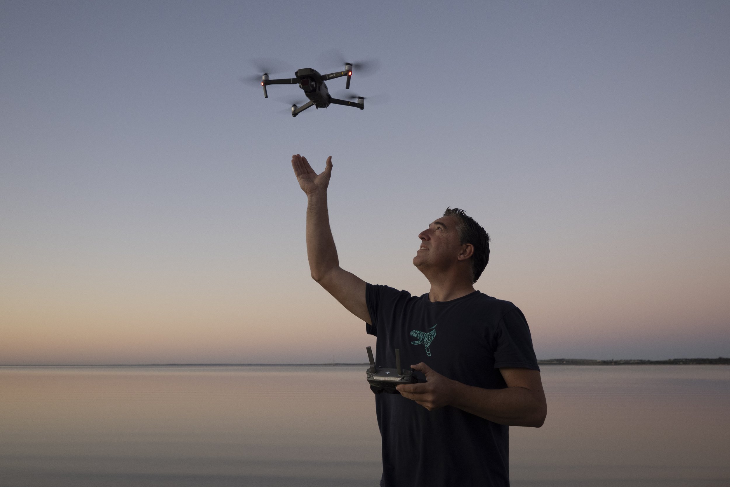 A man standing outdoors at dusk, flying a drone while holding a remote control, with a body of water and a distant shoreline in the background.