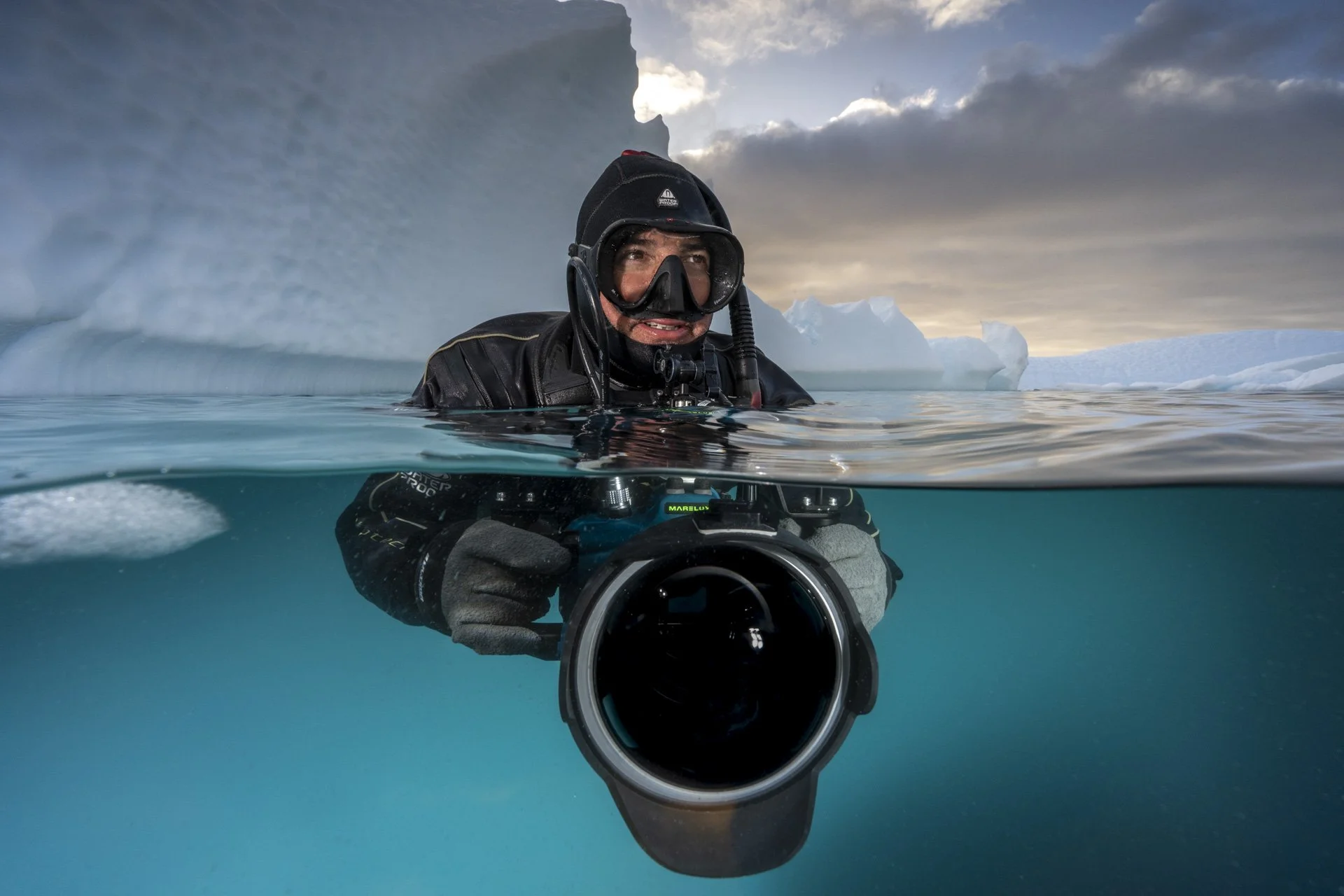 A scuba diver in cold arctic waters taking a photograph, with icebergs and cloudy sky in the background.