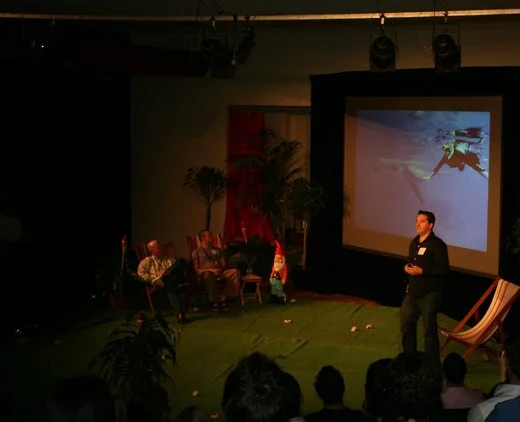 A man is giving a presentation on stage to an audience, with a large screen behind him showing an underwater scene of a diver.