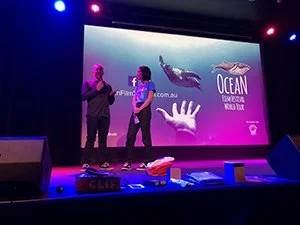 Three people on stage during a presentation or performance, with a large screen behind them displaying a whale image and the text "OCEAN" and "SEABED AND NEAR SHORE." The stage has lights and various equipment visible.