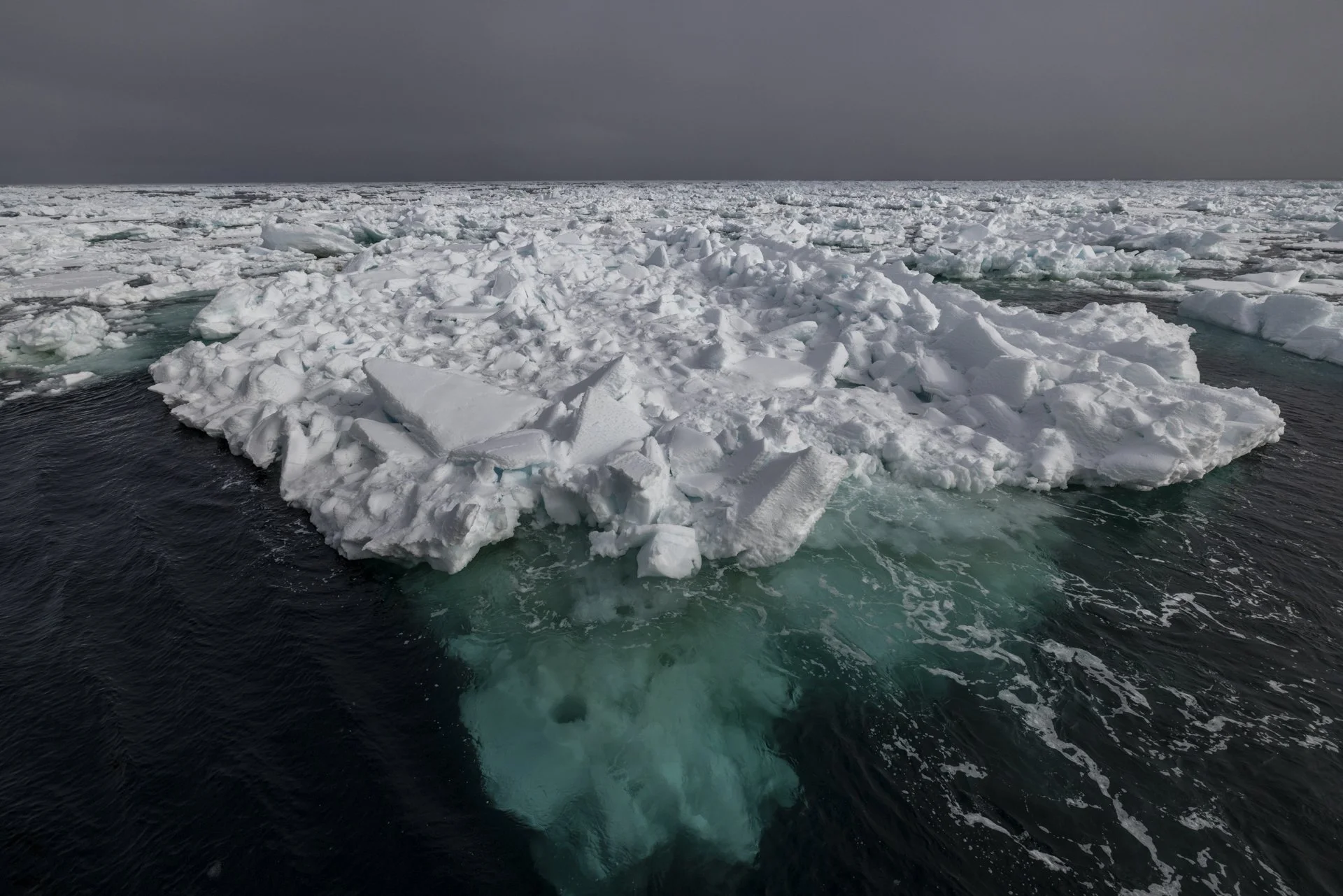 Aerial view of a large ice formation floating in the ocean with dark clouds overhead.