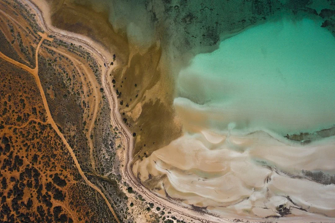 Aerial view of a landscape with a large turquoise body of water, sandy shores, and arid land with dirt roads and sparse vegetation.