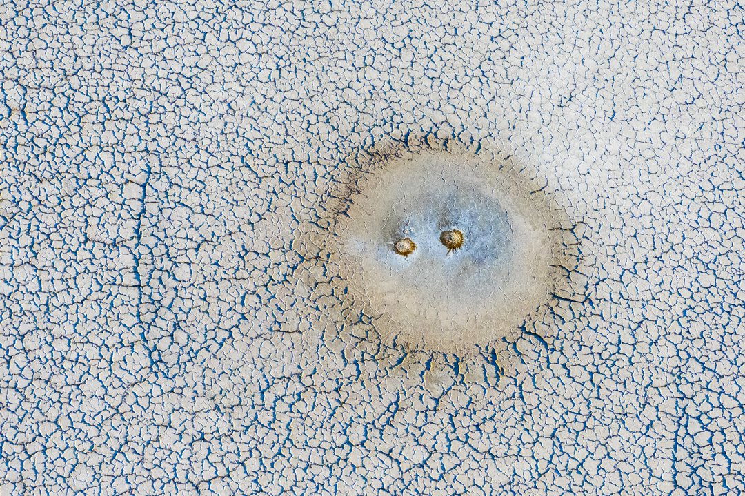 Close-up aerial view of cracked dry earth with a small circular depression in the center, resembling a dried-up salt flat or desert surface.