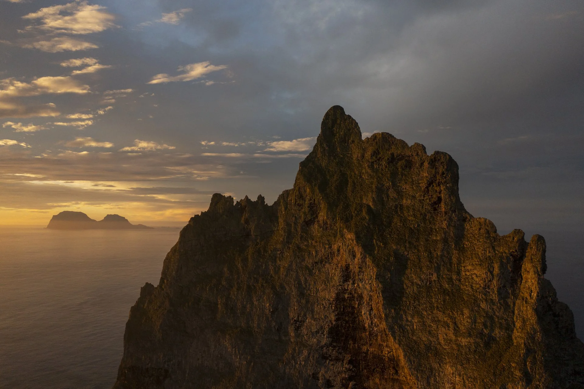 A coastal landscape during sunset with a large rocky cliff in the foreground and distant islands on the horizon under a partly cloudy sky.