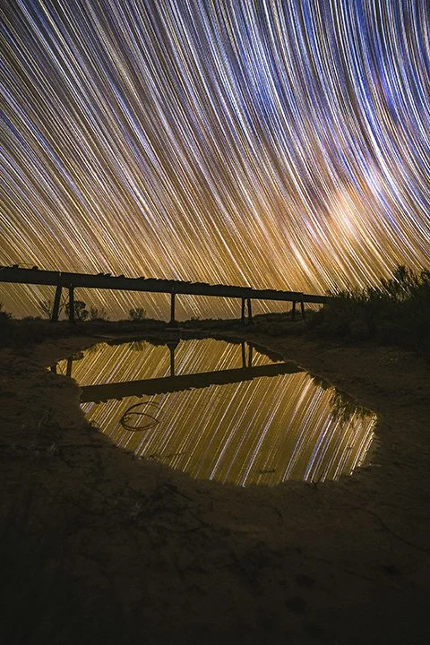Long exposure photo of star trails in the night sky above a landscape with a small pond reflecting the stars and a silhouette of a railroad bridge.