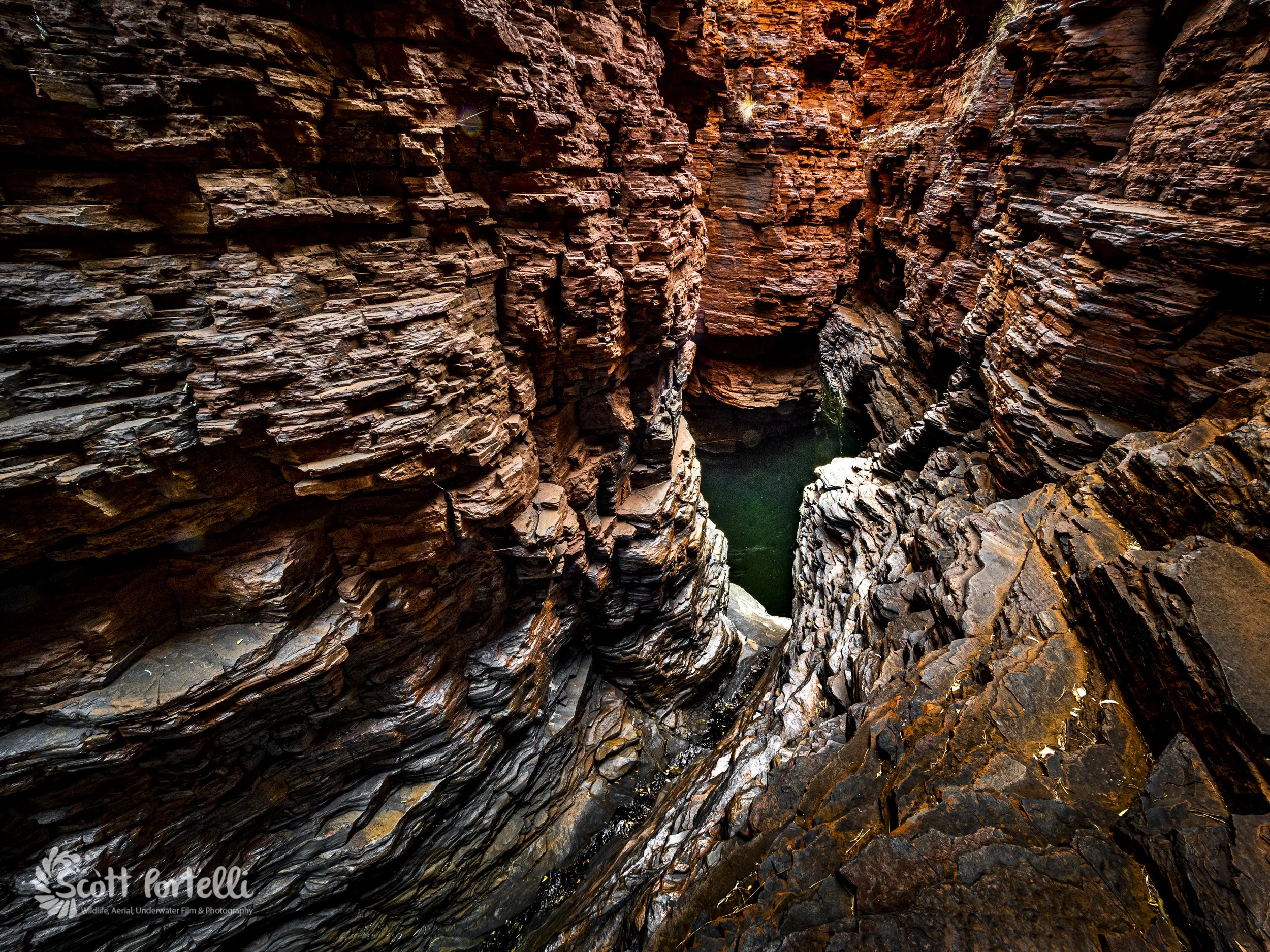 Deep, narrow canyon with layered, reddish-brown rocks on both sides and a small body of water at the bottom.