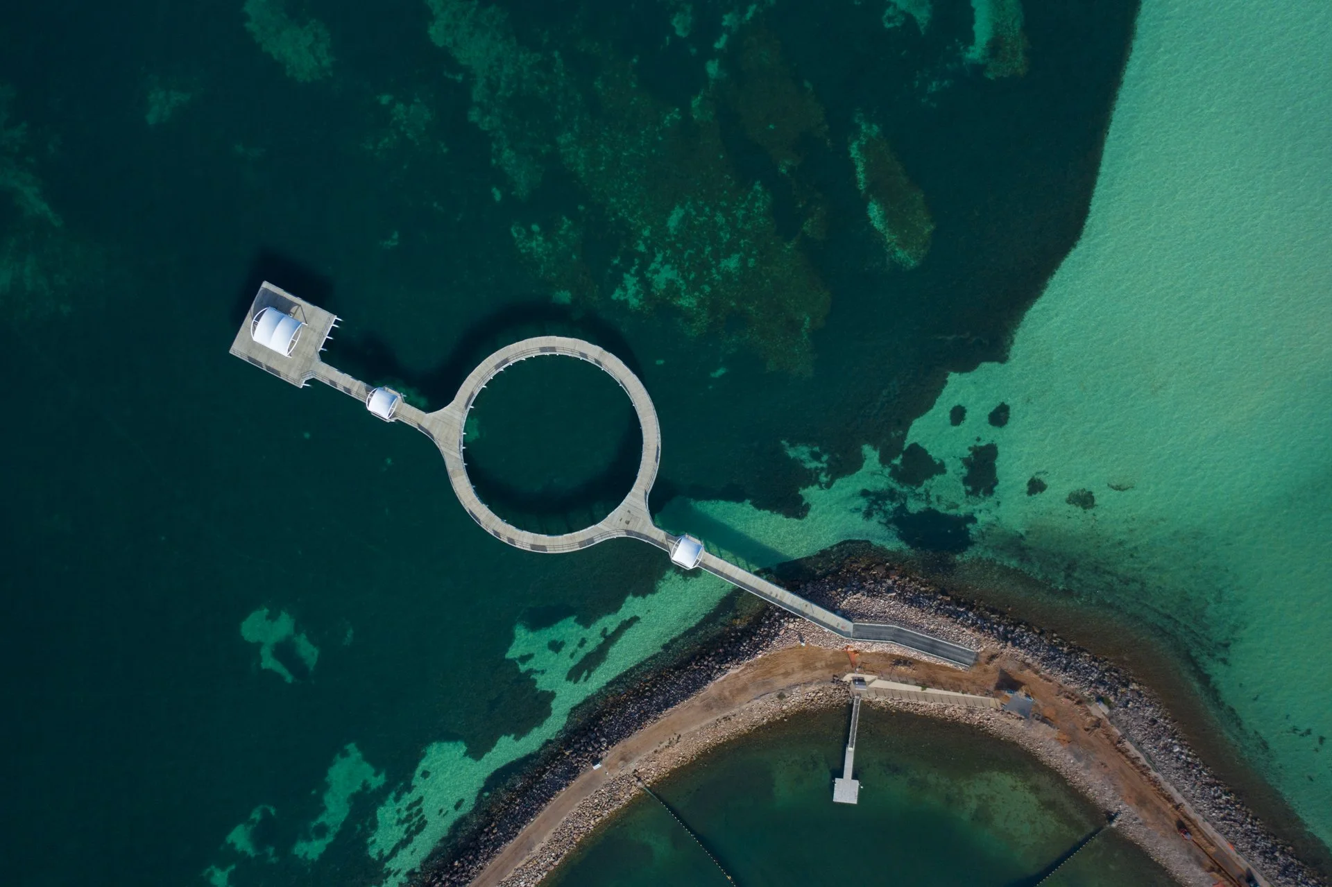An aerial view of a circular pier extending into a body of water with a building on it, positioned along a narrow land strip that curves along the coastline. The water is clear, revealing the seabed with some dark patches.