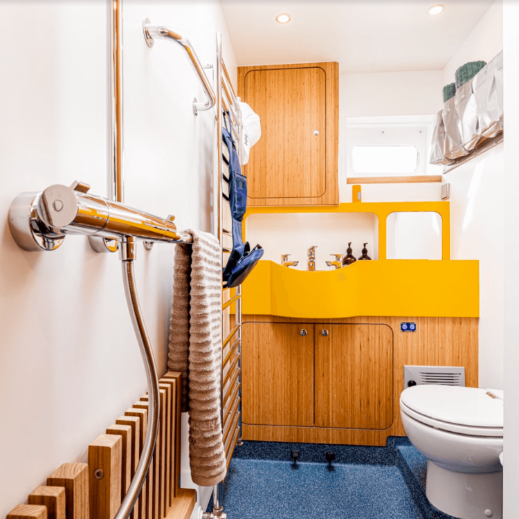 Small bathroom with a yellow sink, wooden cabinet, and a white toilet. Includes a heated towel rack with a towel, and a showerhead on the wall.