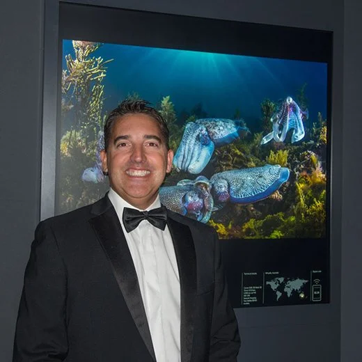 Man in tuxedo standing in front of an underwater-themed digital display with fish and coral.
