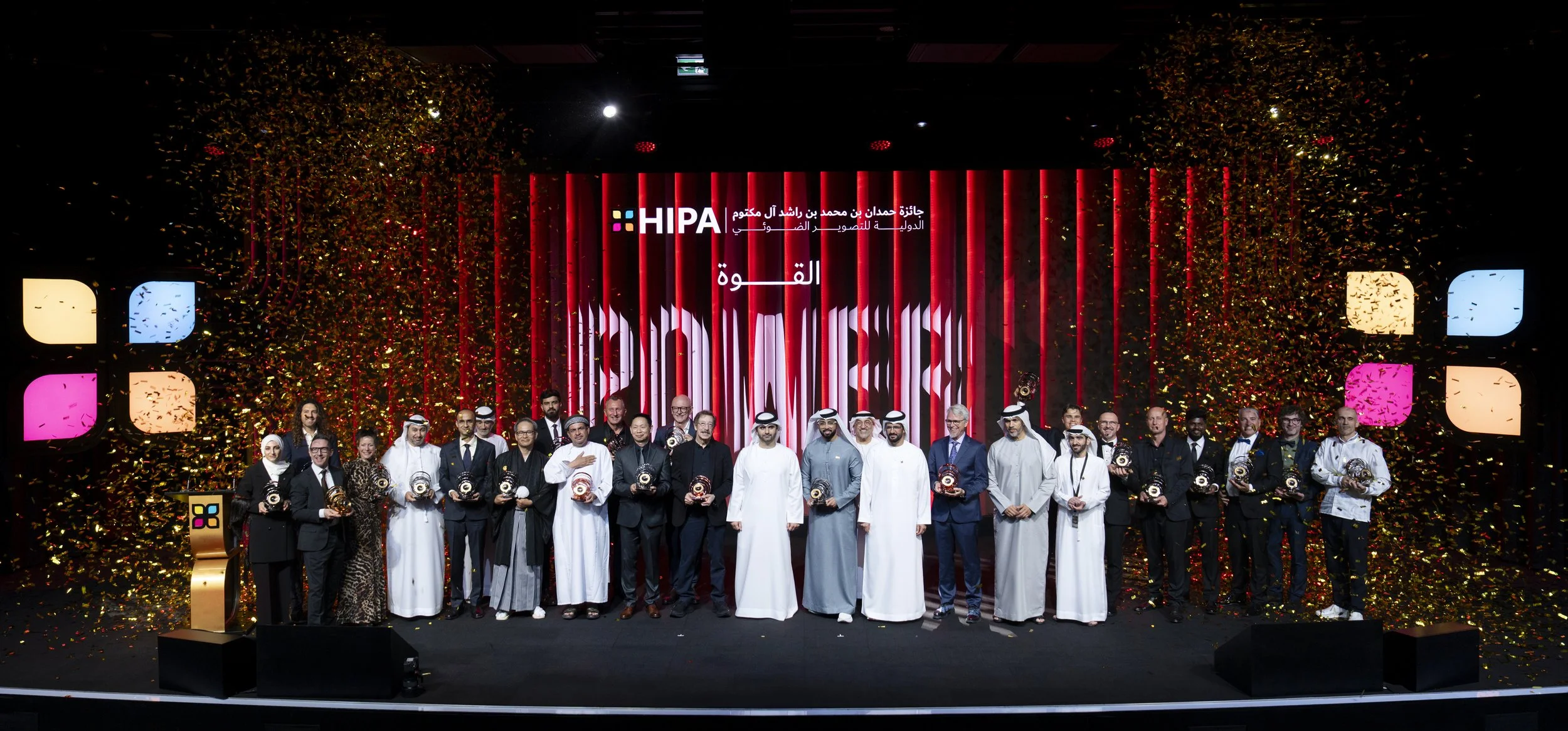 Group of people on stage at an award ceremony, holding trophies, with confetti falling and a large screen displaying Arabic text and the logo of the event.