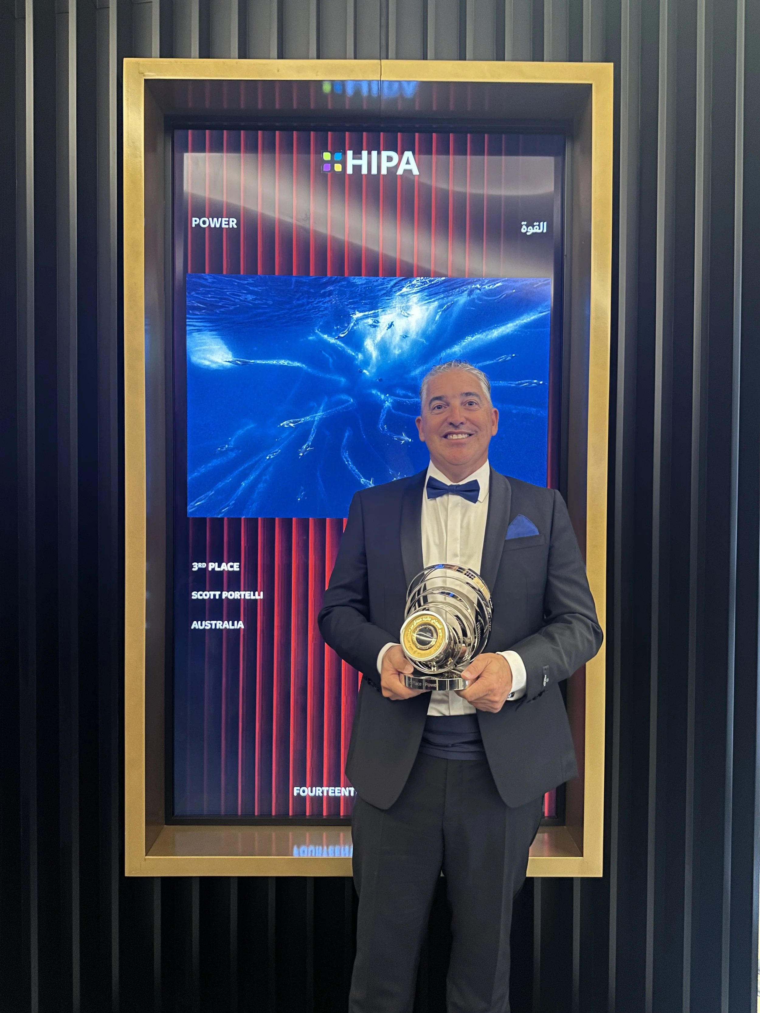 A man in a black tuxedo and bow tie holding a large silver trophy in front of a digital display with the text 'HIPA' and 'Power' on it, at an awards event.