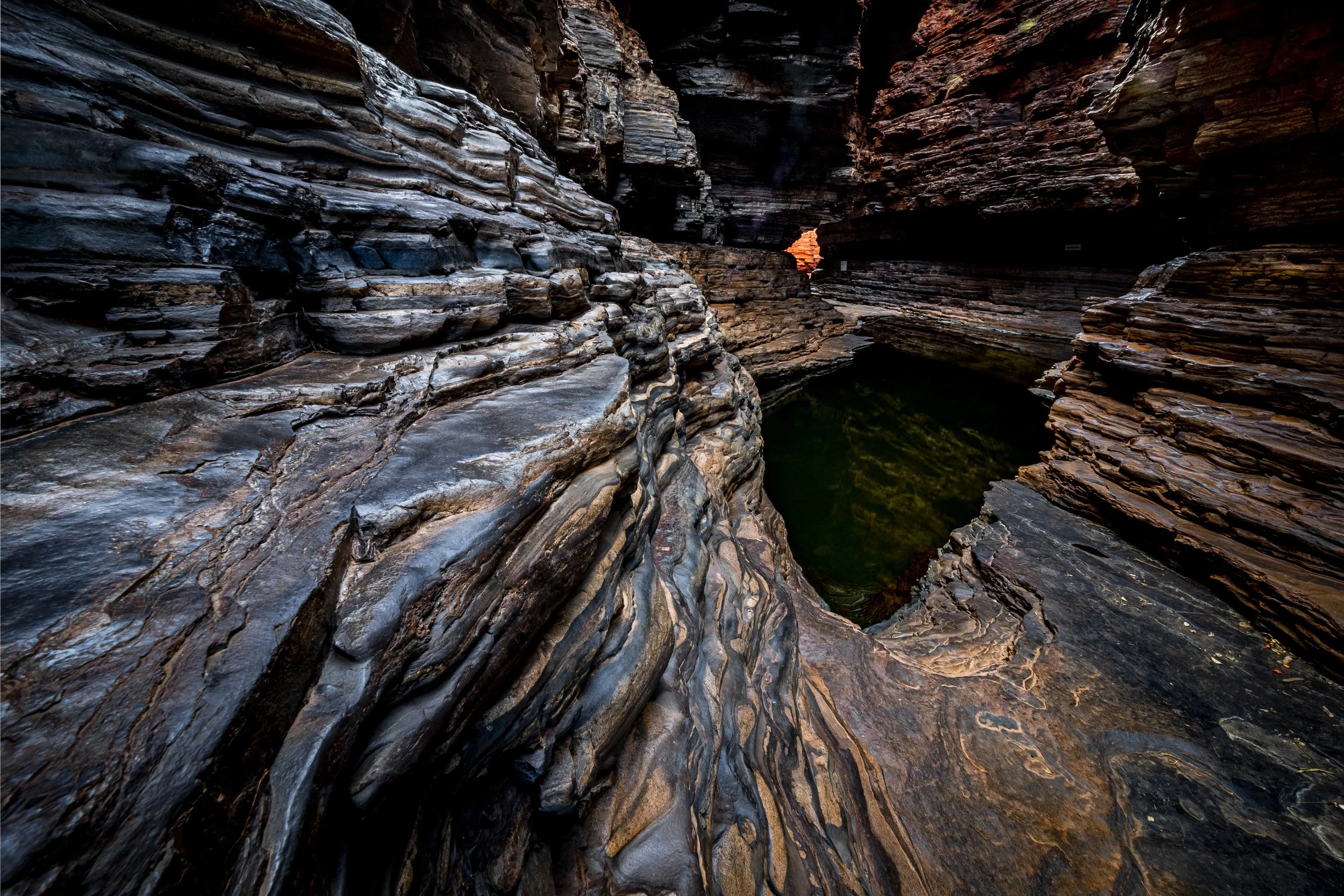 A deep, narrow canyon with layered rock formations in dark shades of brown, black, and gray; a small patch of water at the bottom reflecting some light, and a faint glow of orange visible through a small opening in the distance.