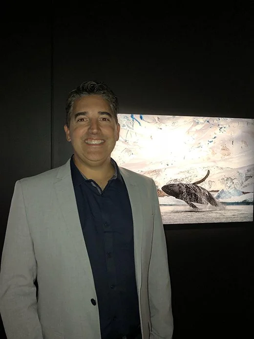 Scott Portelli wearing a light gray blazer and navy shirt, smiling while standing in front of an aquarium display with a whale in icy waters.