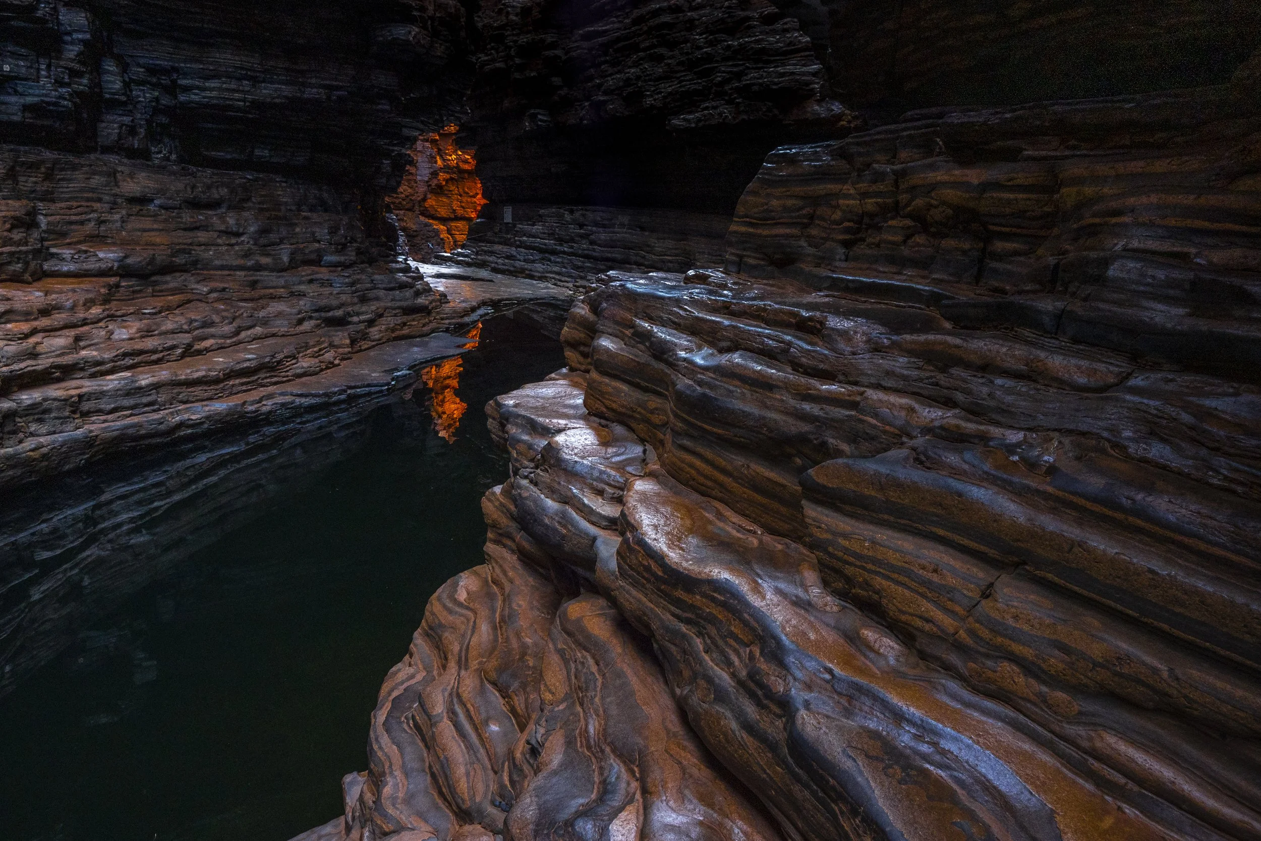 View of a dark, narrow canyon with layered rock walls and a small pool of water reflecting orange light from an opening.