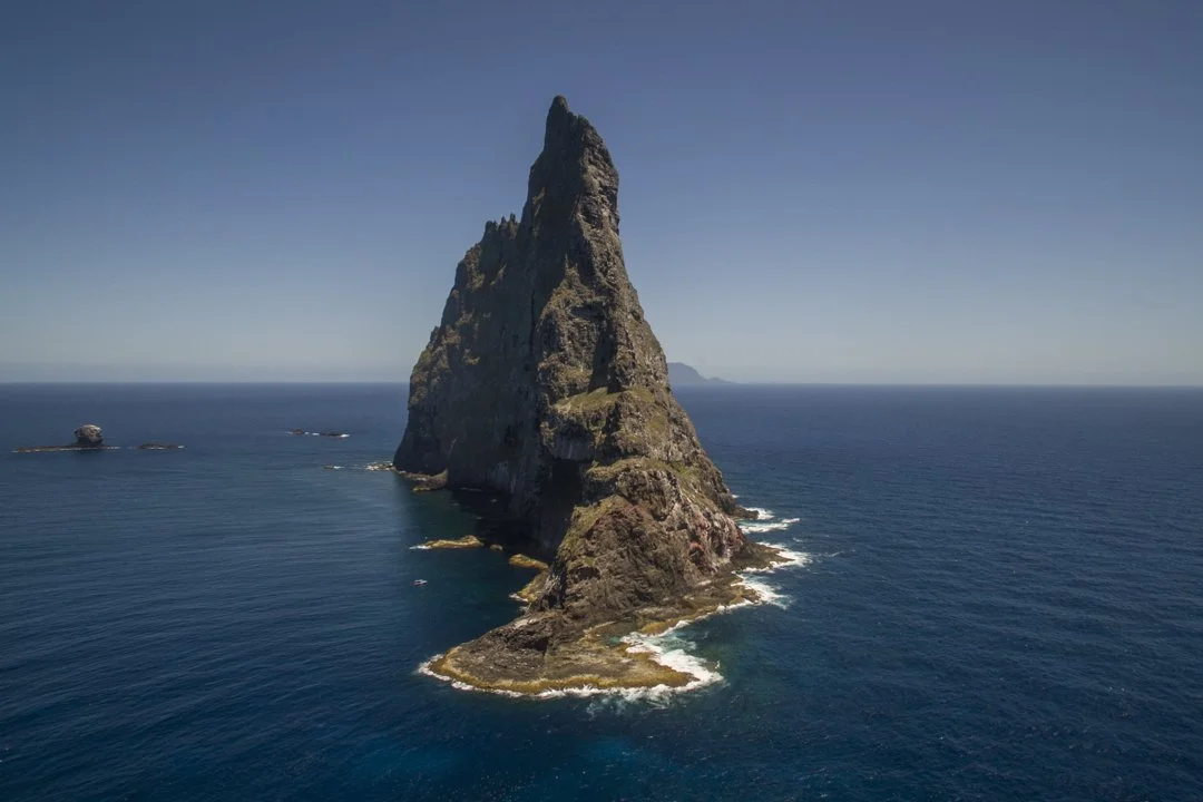 A large rocky sea stack rising from the ocean with smaller rocks around it, under a clear blue sky.