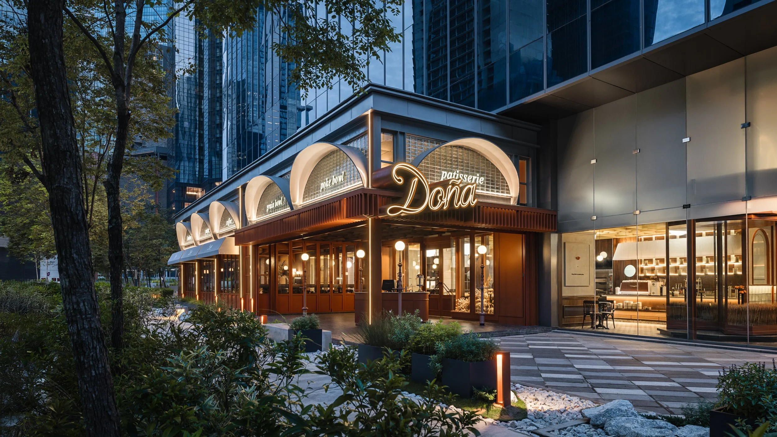 A modern city street scene at dusk featuring a restaurant called 'Dona' with illuminated signage, large glass windows, and a stylish exterior. Tall skyscrapers with reflective glass facades and trees line the street.