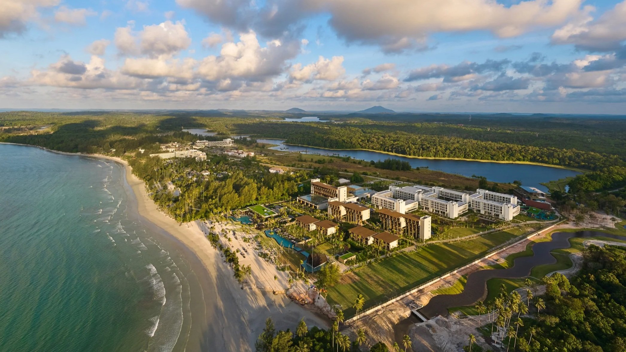 Aerial view of a coastal resort with a sandy beach, turquoise ocean, palm trees, and modern buildings, surrounded by green hills and mountains under partly cloudy skies.