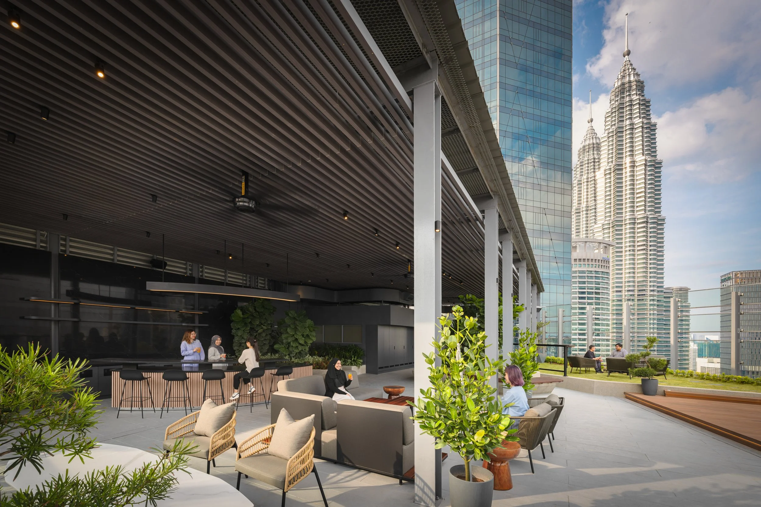People socializing and relaxing on a rooftop terrace with modern furniture and greenery, overlooking tall skyscrapers and the city skyline.