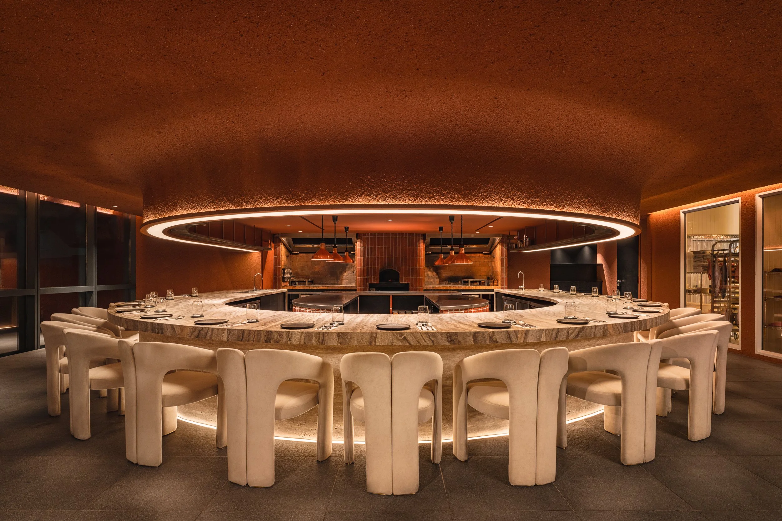Modern restaurant interior with a large circular marble dining table surrounded by white chairs, featuring pendant lighting and a brick oven in the background.