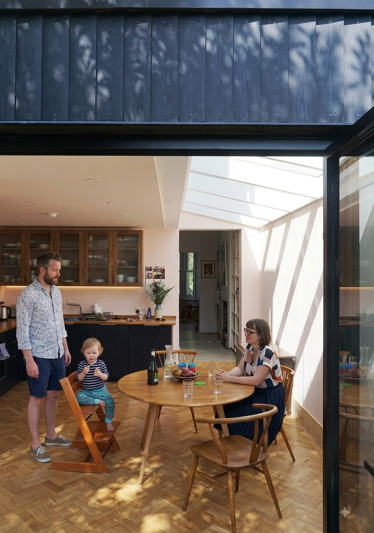 Family gathering in a modern, sunlit kitchen and dining area with wooden flooring, a round table, and three people: a man, a woman, and a small child.