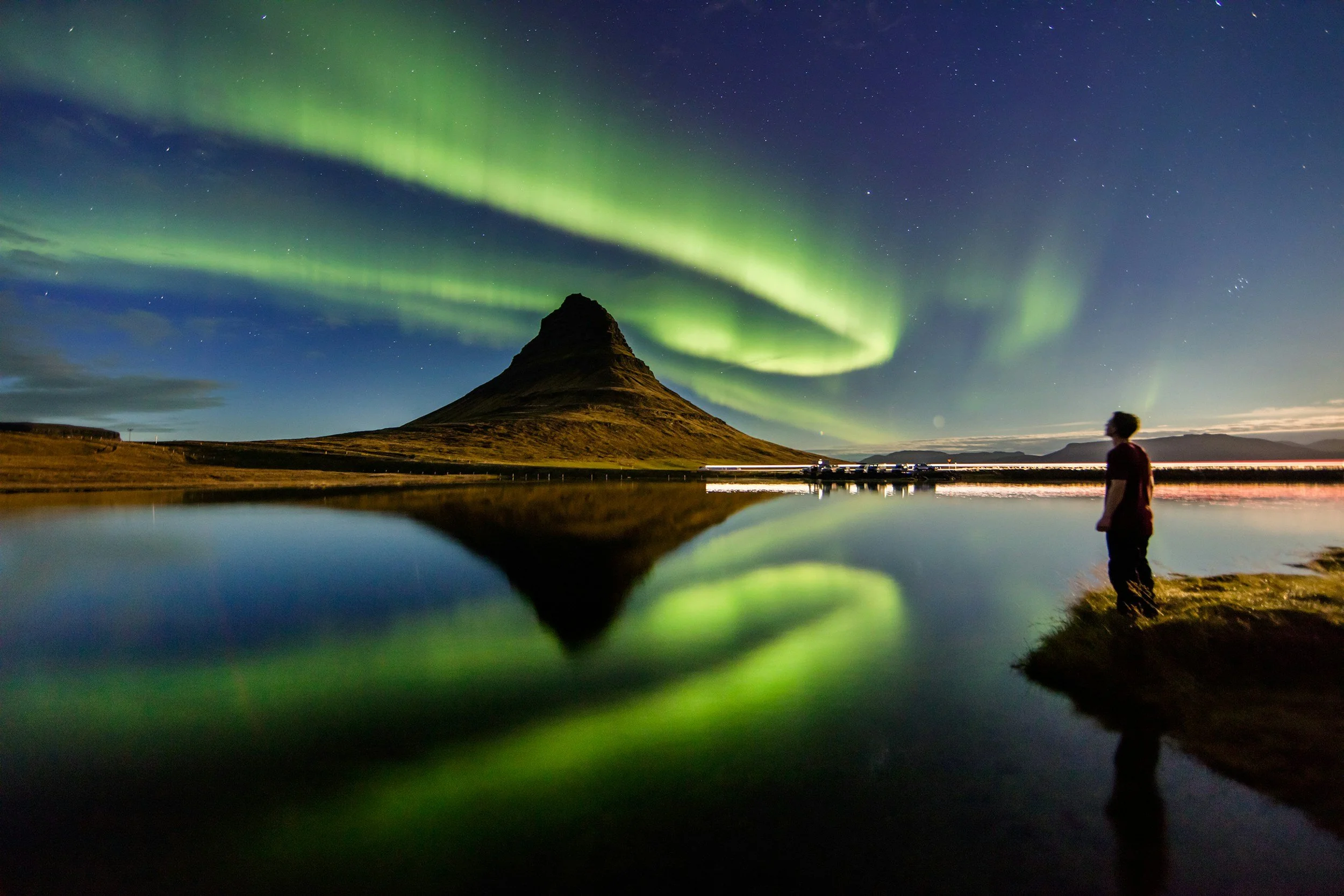 Nighttime landscape with a person standing by water, observing the Northern Lights over a mountain, with stars in the sky.