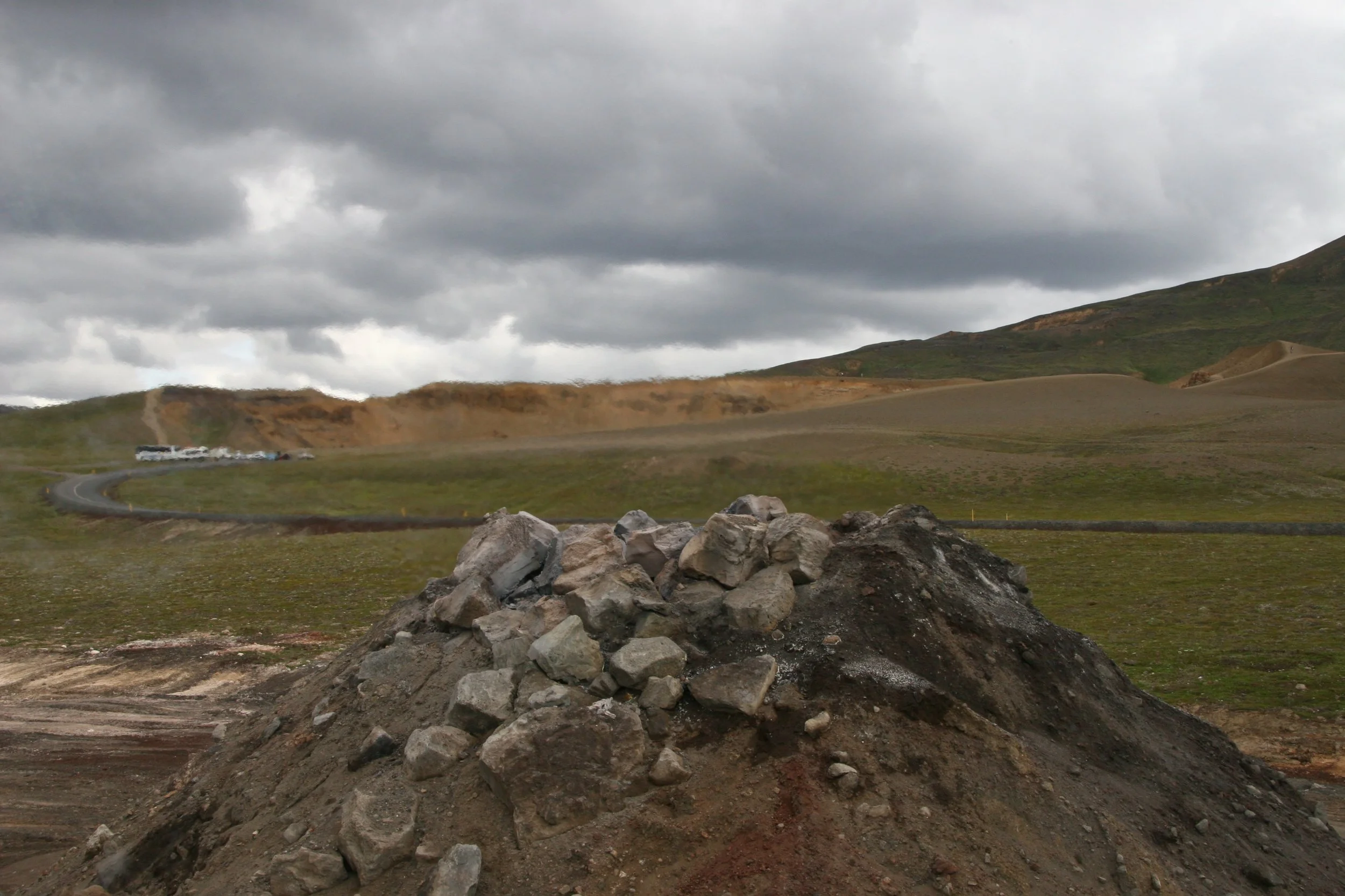 A mountain landscape under a cloudy sky with a winding road, green hills, and a pile of rocks in the foreground.