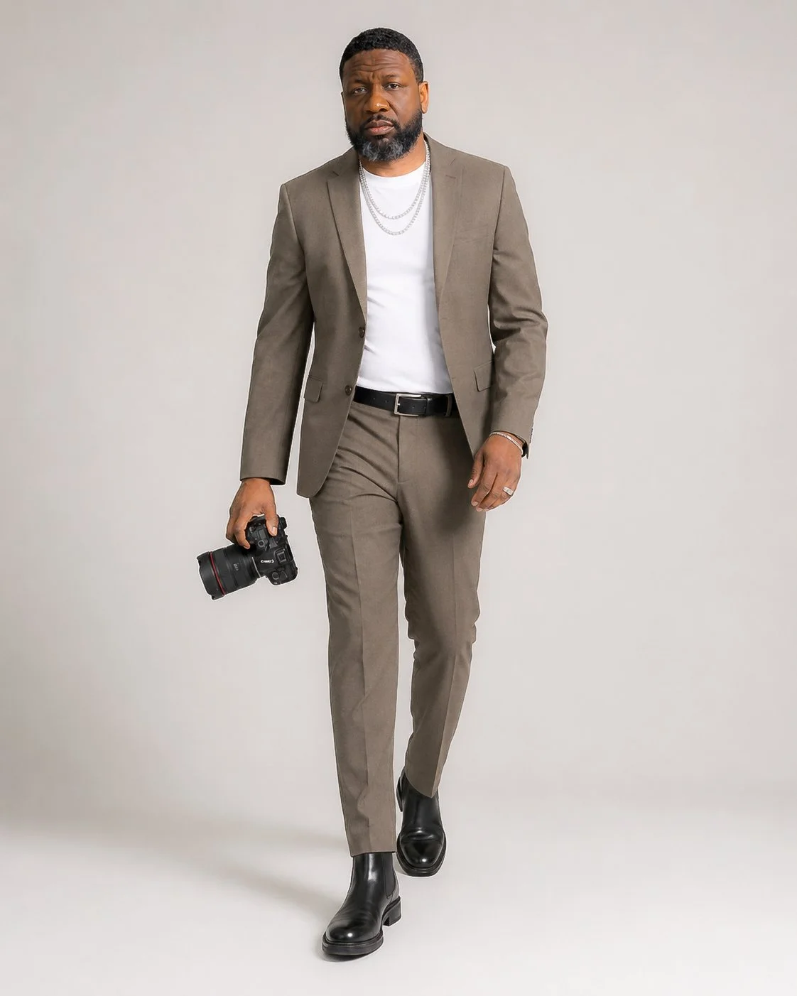 A man in a taupe suit with a white t-shirt, holding a professional camera, walking against a plain light background.