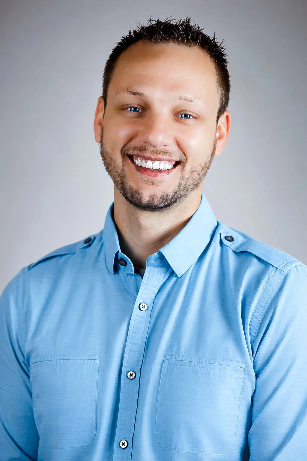 Corporate business headshot. A smiling man with short dark hair, blue eyes, and beard wearing a light blue button-up shirt against a neutral gray background.