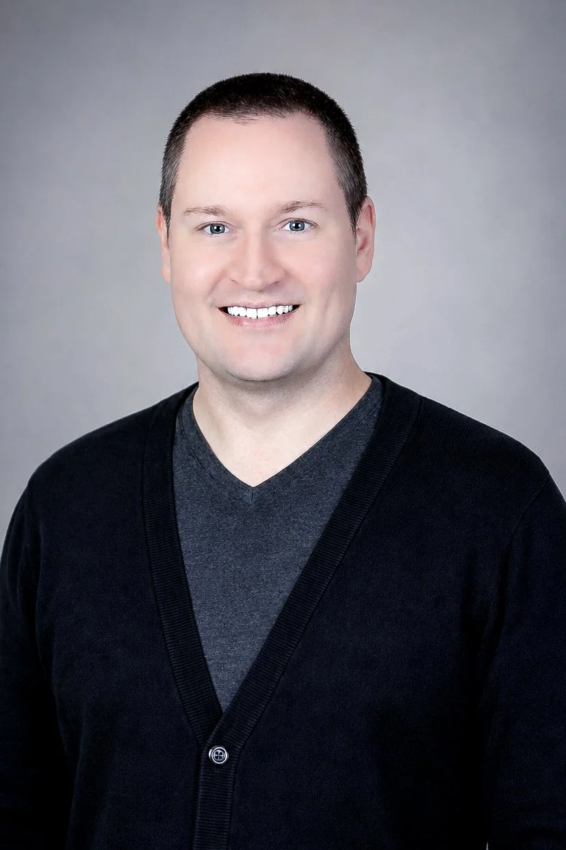 Corporate headshot. Portrait of a smiling man with short dark hair, wearing a dark gray V-neck shirt underneath a black cardigan, against a plain gray background.