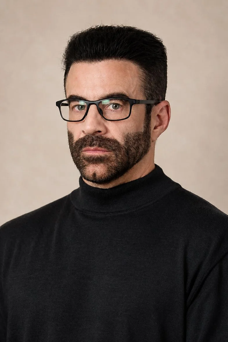Corporate Headshot. Man with dark hair, beard, glasses, wearing a black turtleneck, looking at the camera, neutral background.