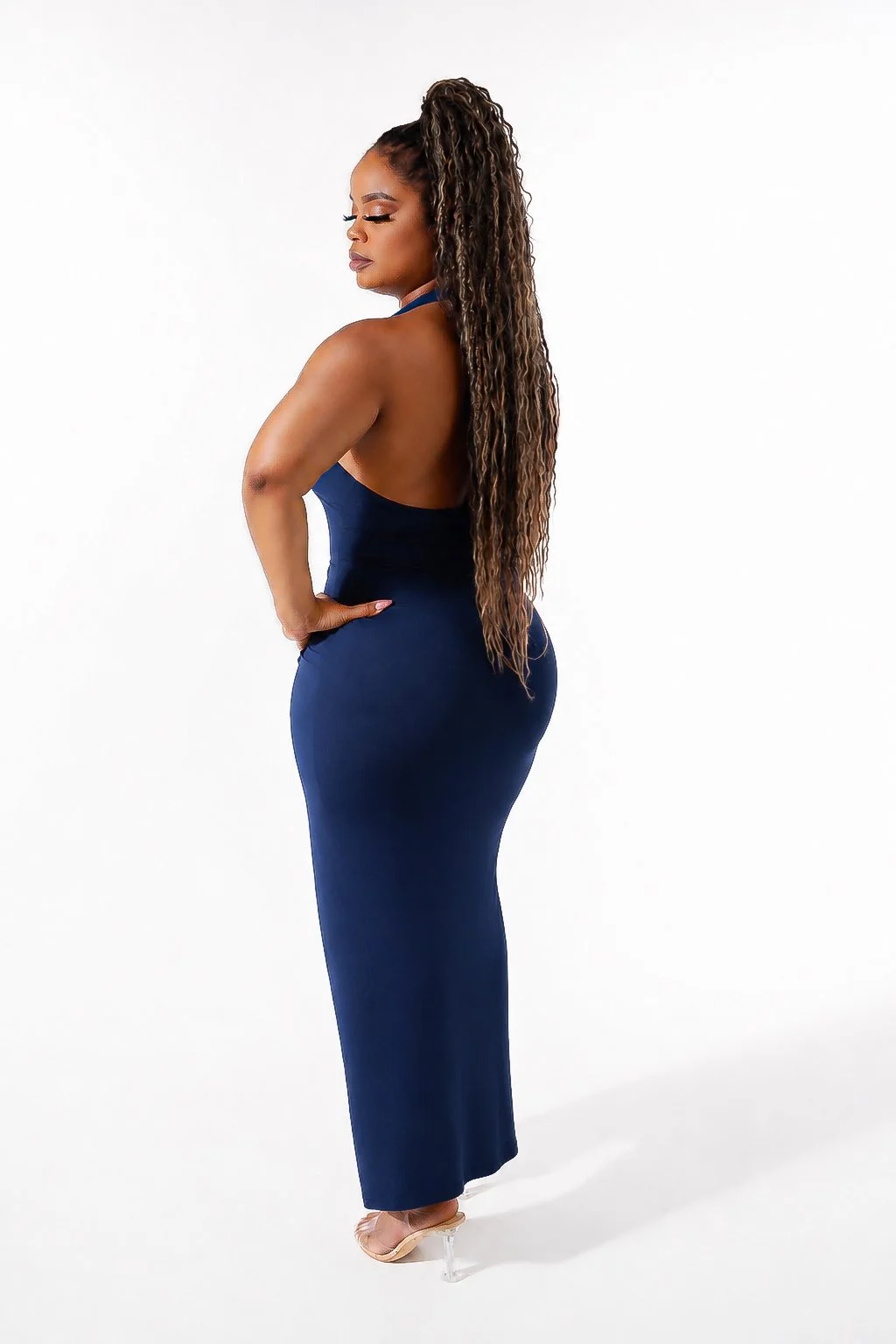 Fashion Photography. A woman with long, curly hair in a high ponytail, wearing a fitted navy blue dress and clear high heels, standing sideways against a plain white background.