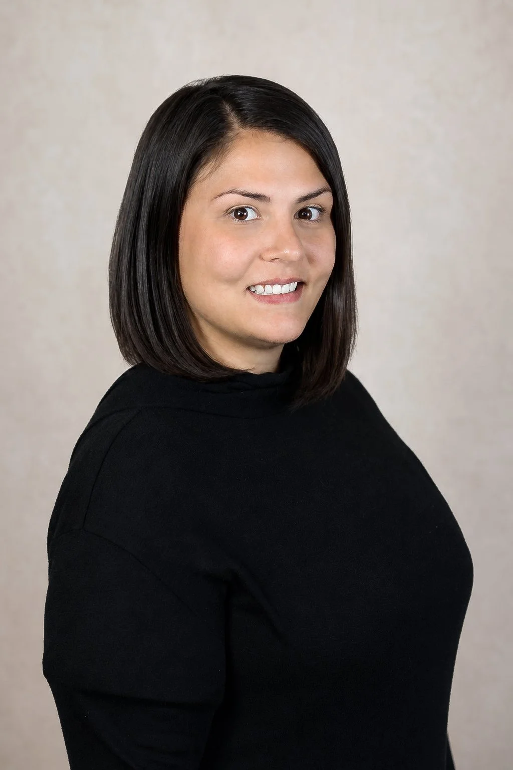 Corporate business headshot. A woman with straight dark brown hair, wearing a black turtleneck, smiling slightly, looking at the camera in front of a plain background.