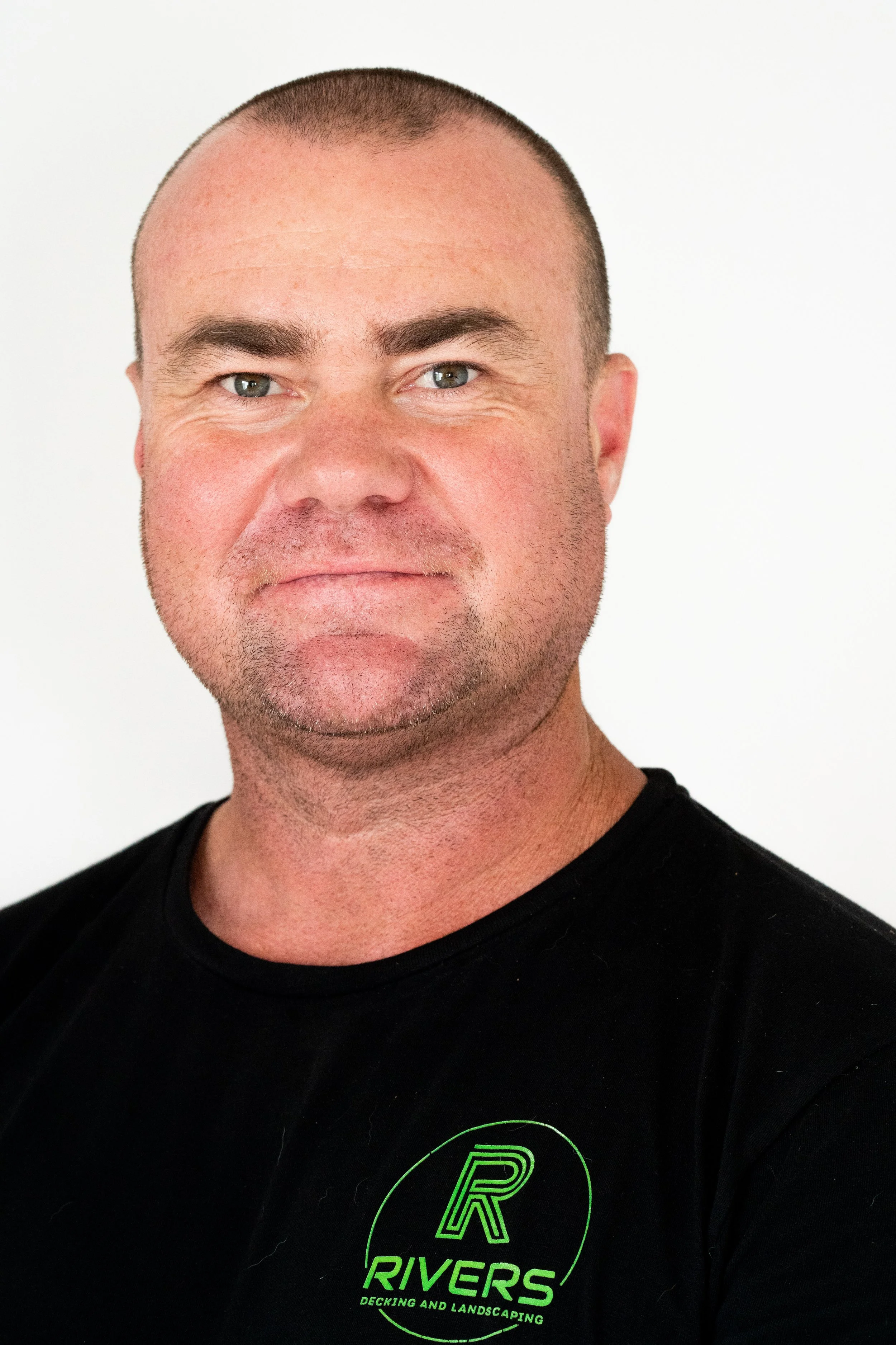 A man with a shaved head and light skin, wearing a black T-shirt with green text that reads "Rivers Decking and Landscaping." The background is plain white.