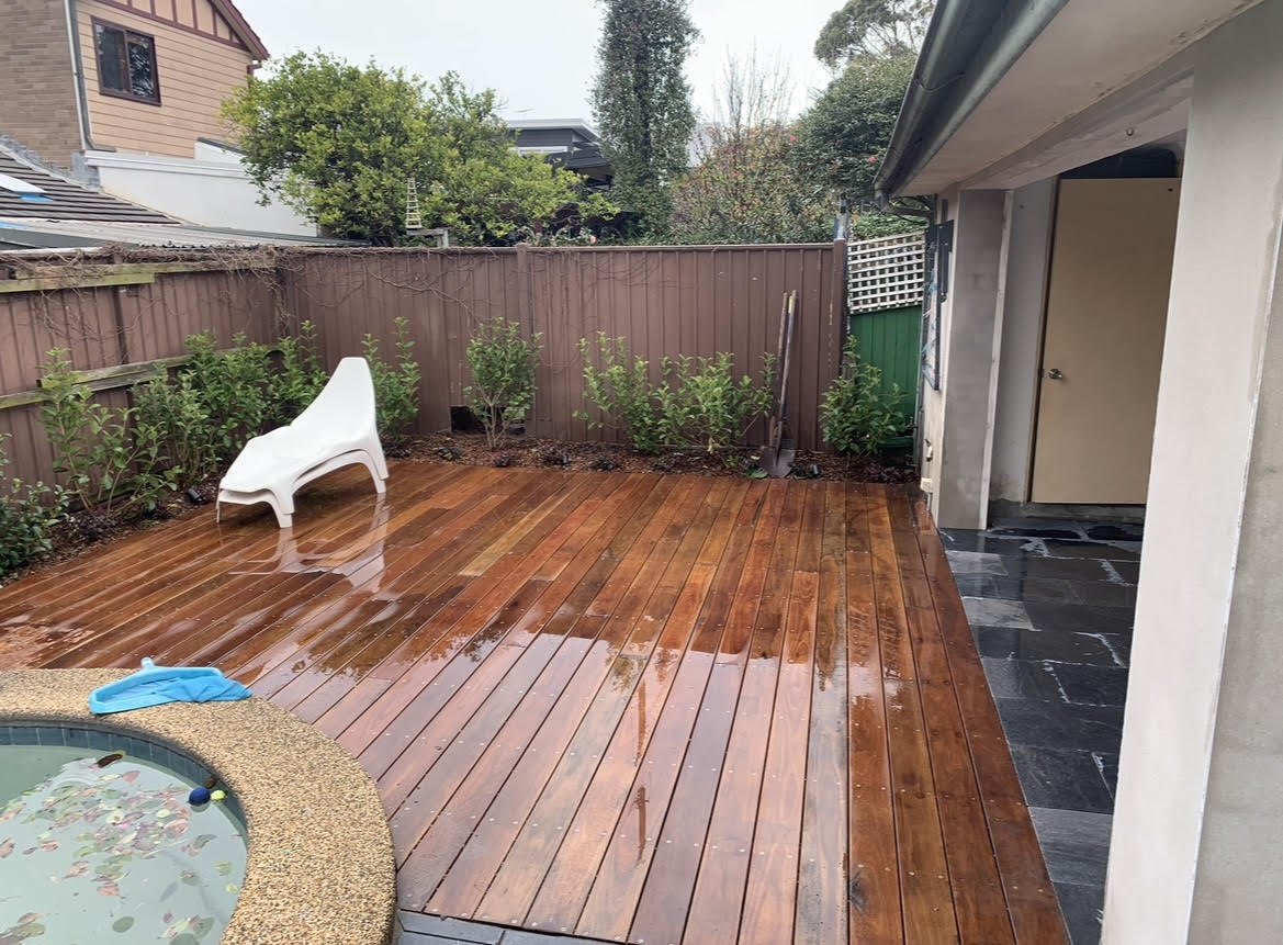 Wet wooden deck with a white chair and small plants along the fence, adjacent to a house with an open door and a small pond with an inflatable blue float in the foreground.