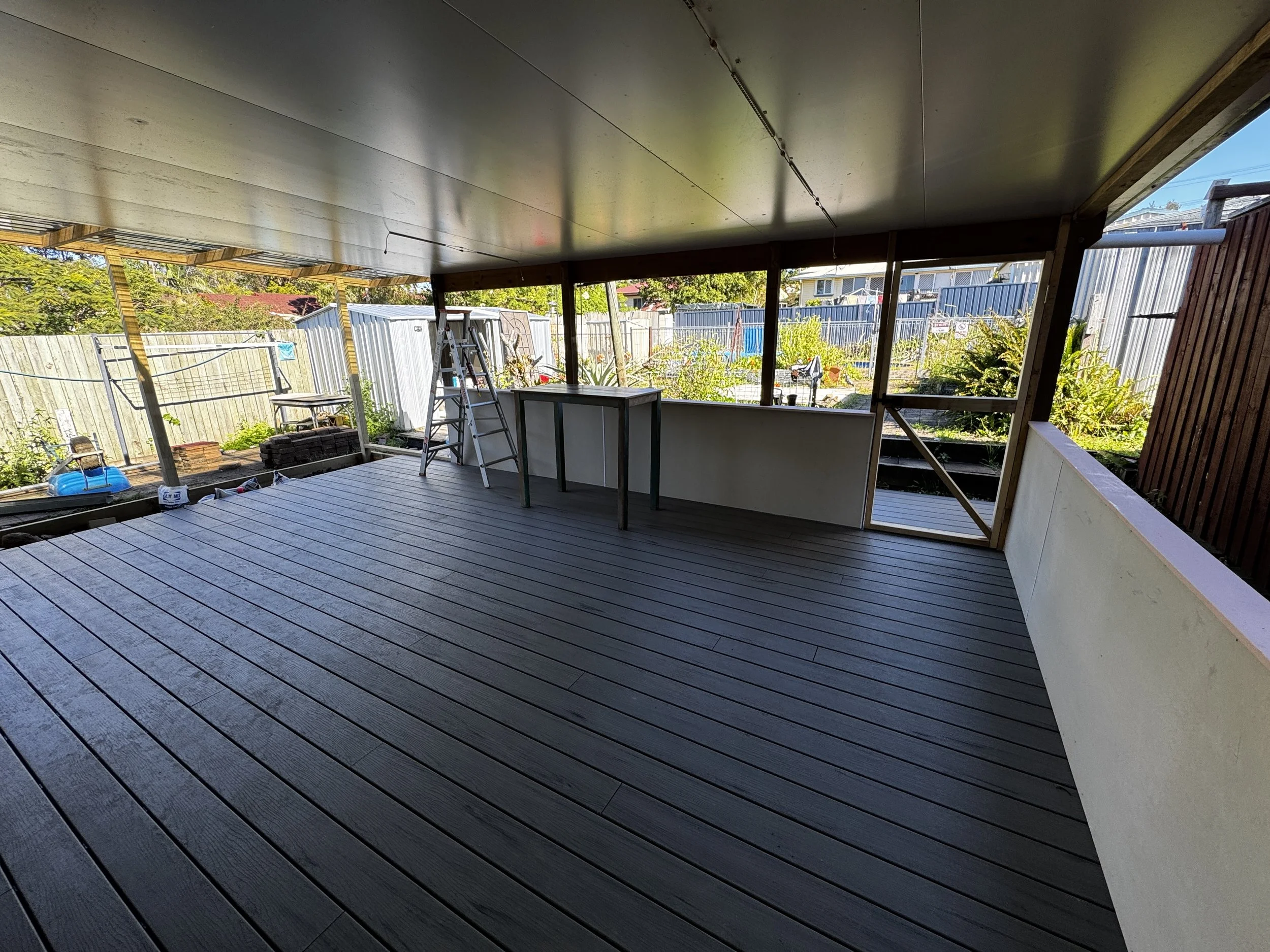 Under construction porch area with a ladder, tools, and building materials on a wooden deck, with a view of a fenced backyard with trees and houses.