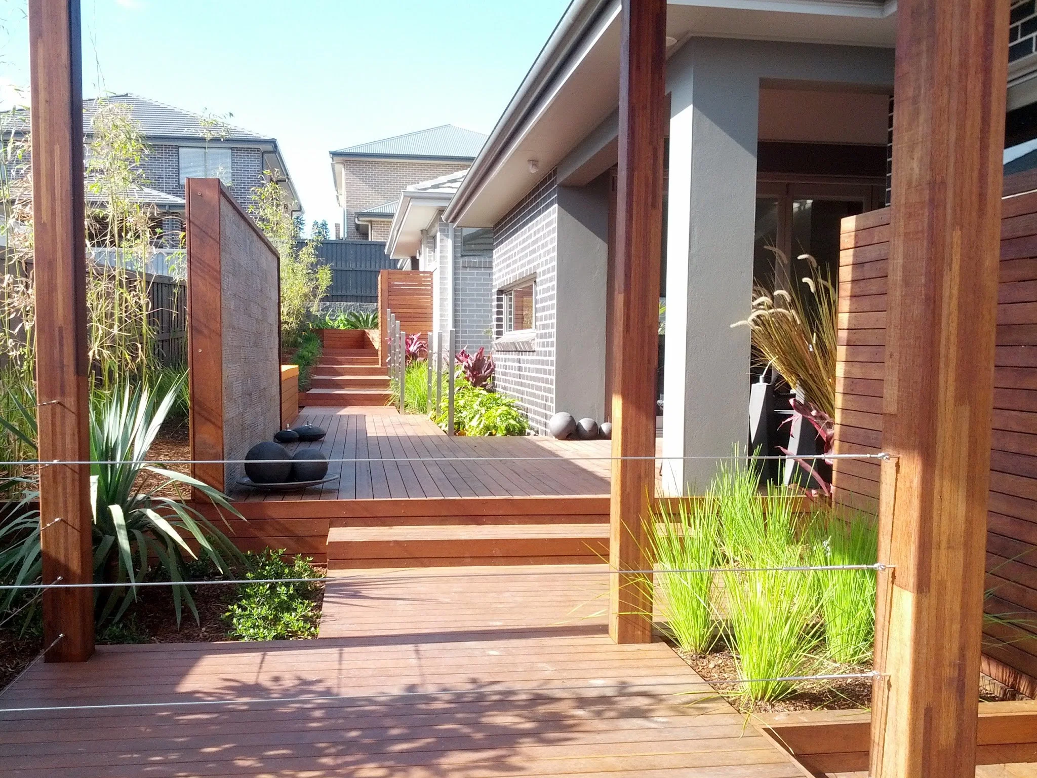 A modern backyard patio with wooden deck, planters, and a staircase leading to a garden area, surrounded by a fence and neighboring houses.