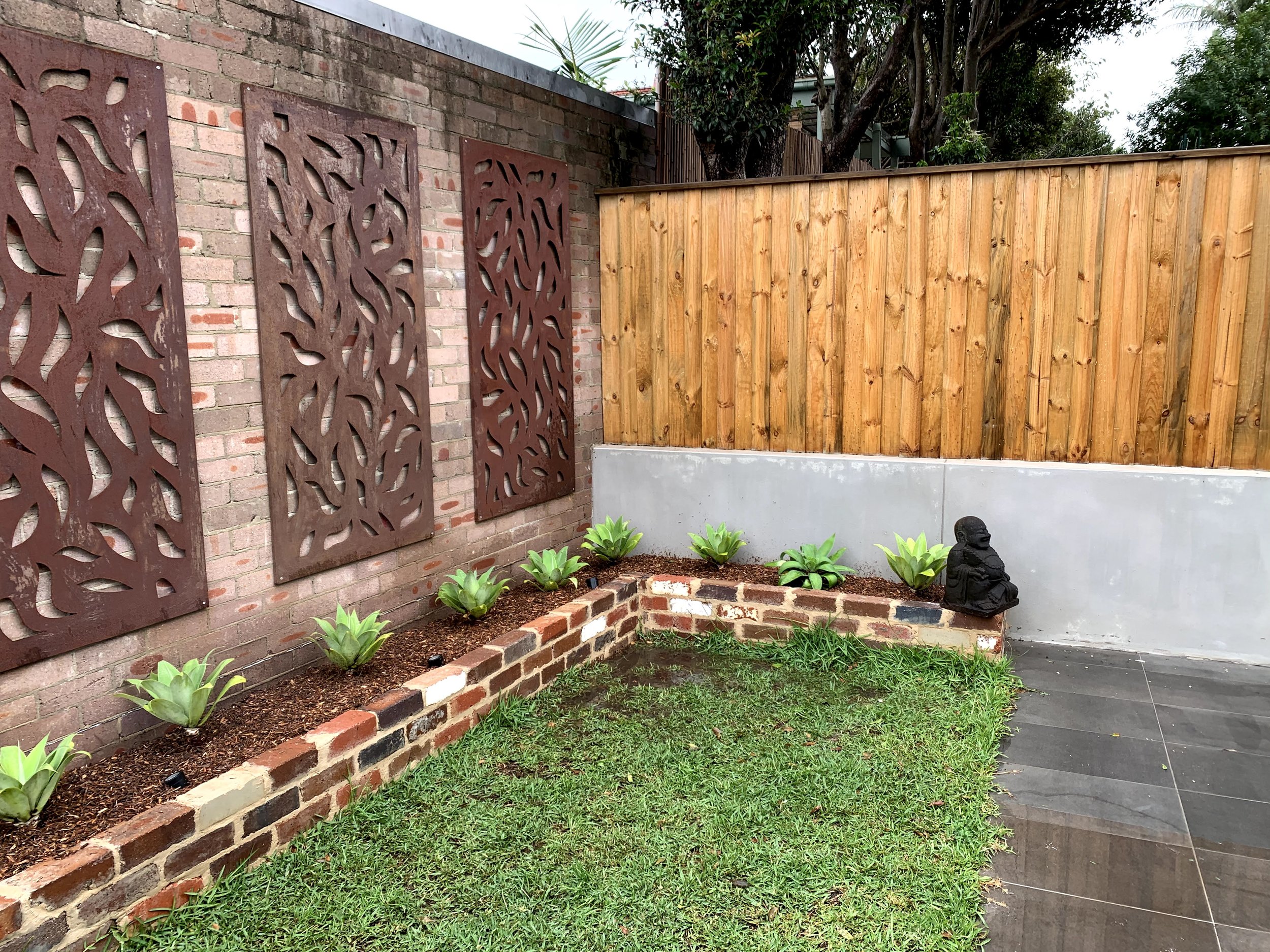 A backyard garden with a decorative brick wall, metal art panels, a garden bed with green plants, and a small statue of a person seated in a relaxed pose. There is a wooden privacy fence and trees in the background.