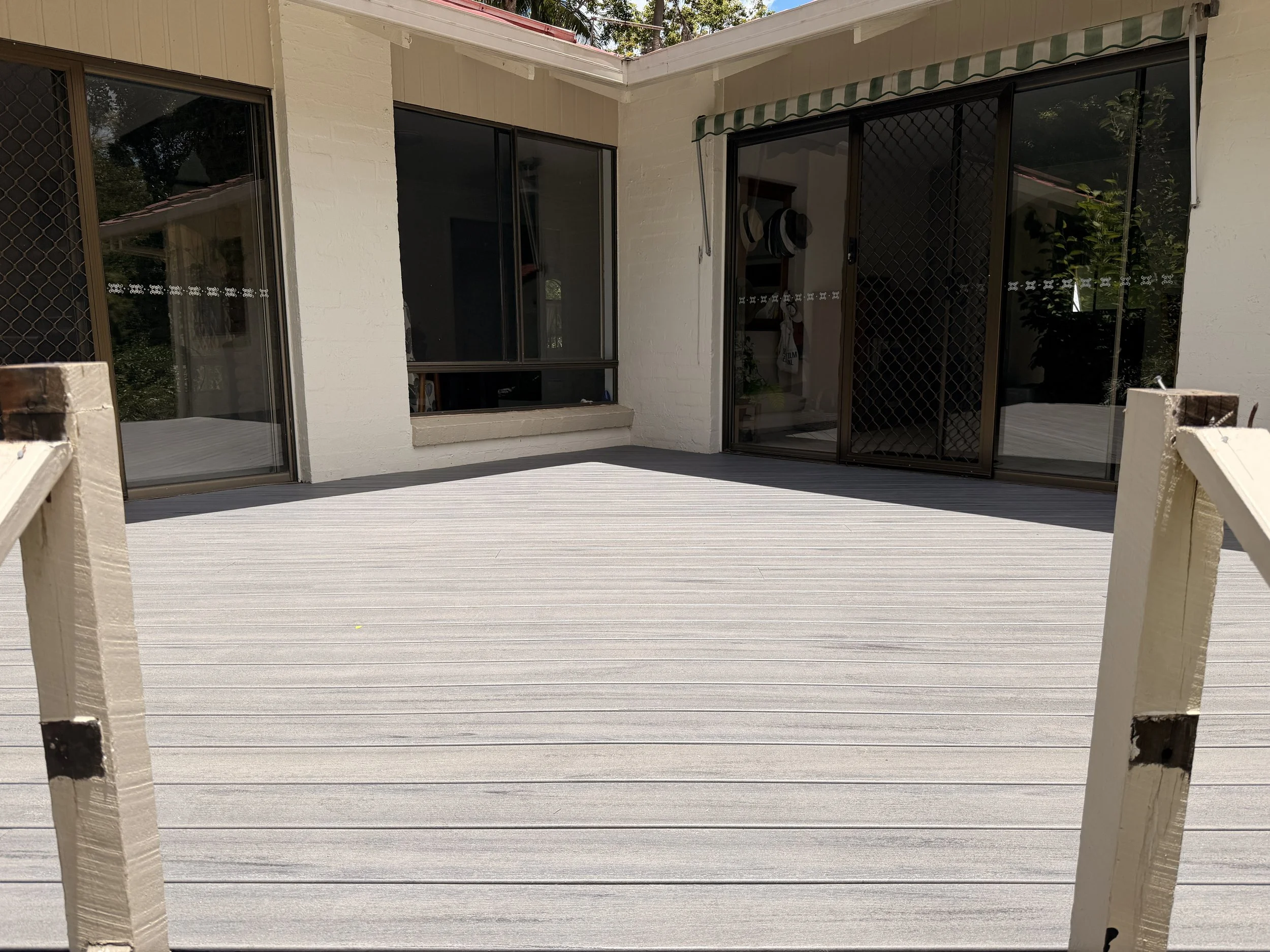 A beige deck on the exterior of a house, with three large sliding glass doors. One door is closed, another is partially open, and a third is fully open. There is a small ledge in front of the windows, with two hats hanging on hooks inside. The house 