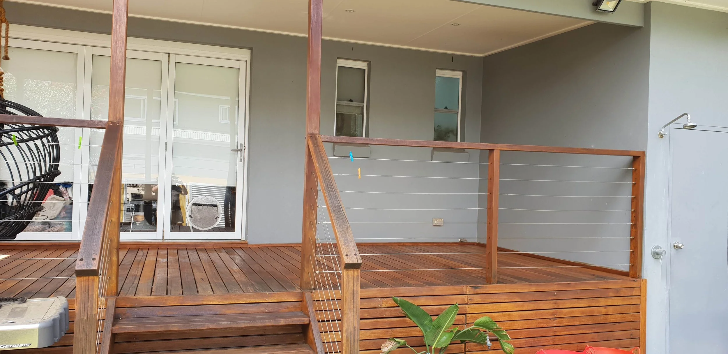 View of a wooden deck with a railing, three windows, a glass door, and a wall-mounted outdoor shower head. There is a plant with large green leaves in the foreground and some outdoor items visible through the glass door.