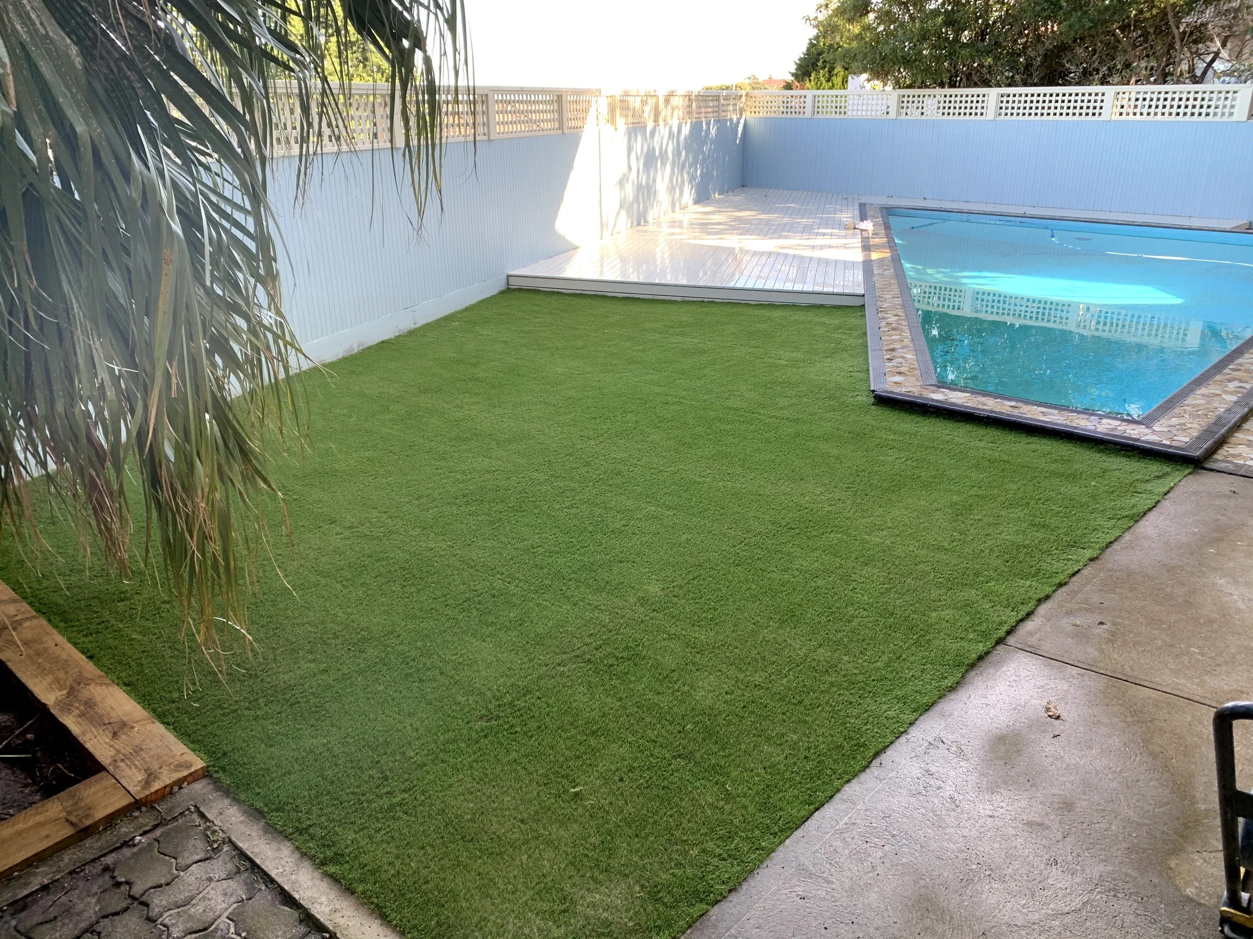 A backyard with a green artificial grass lawn, a small pool with a tiled border, a white wooden fence, and some trees and shrubbery in the background.