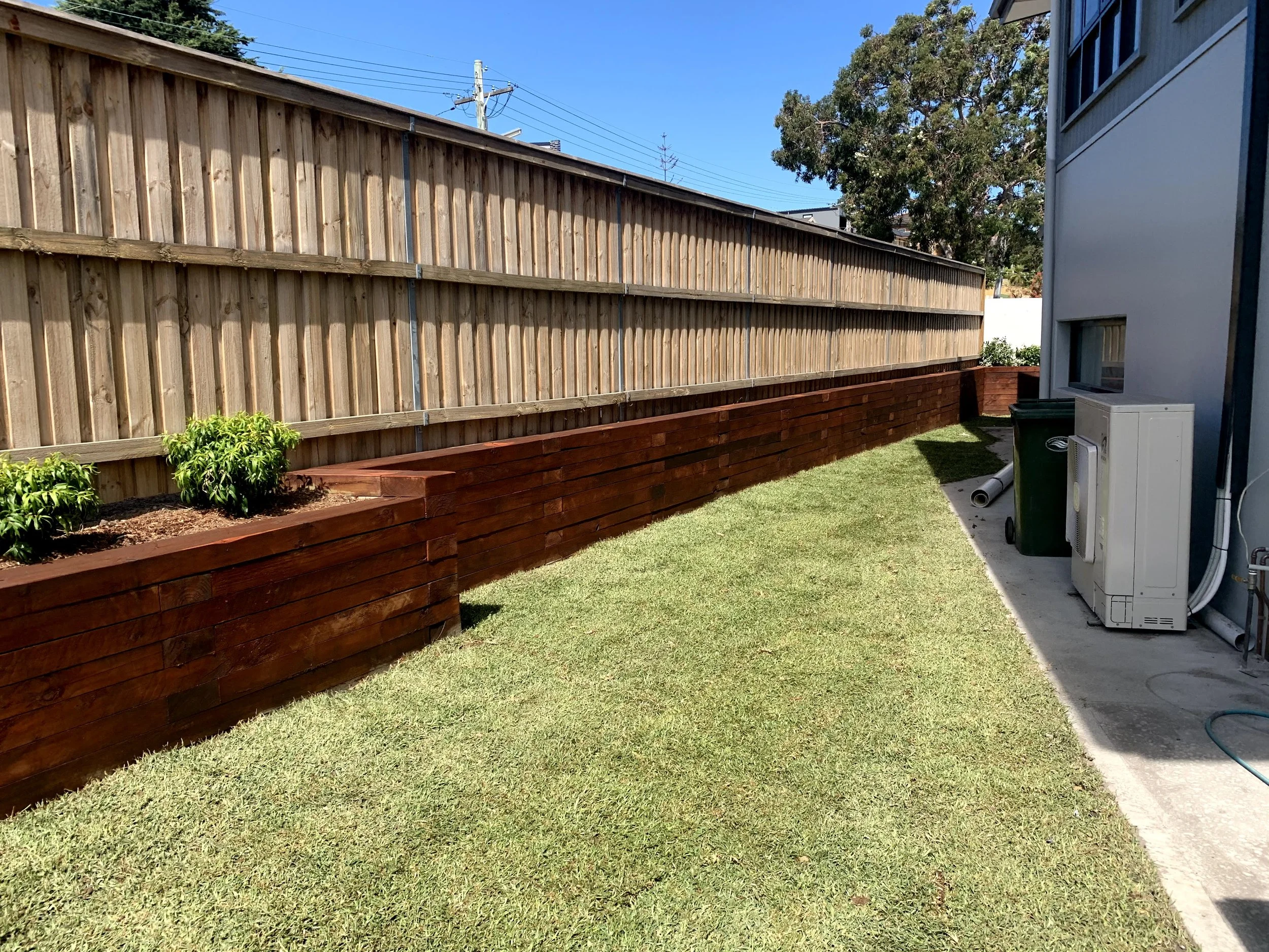 A backyard with a tall wooden privacy fence, green grass, and a few small bushes in a wooden planter. On the right side, part of a house with a window and a utility box is visible. The sky is clear and blue, with utility lines and a tree in the backg