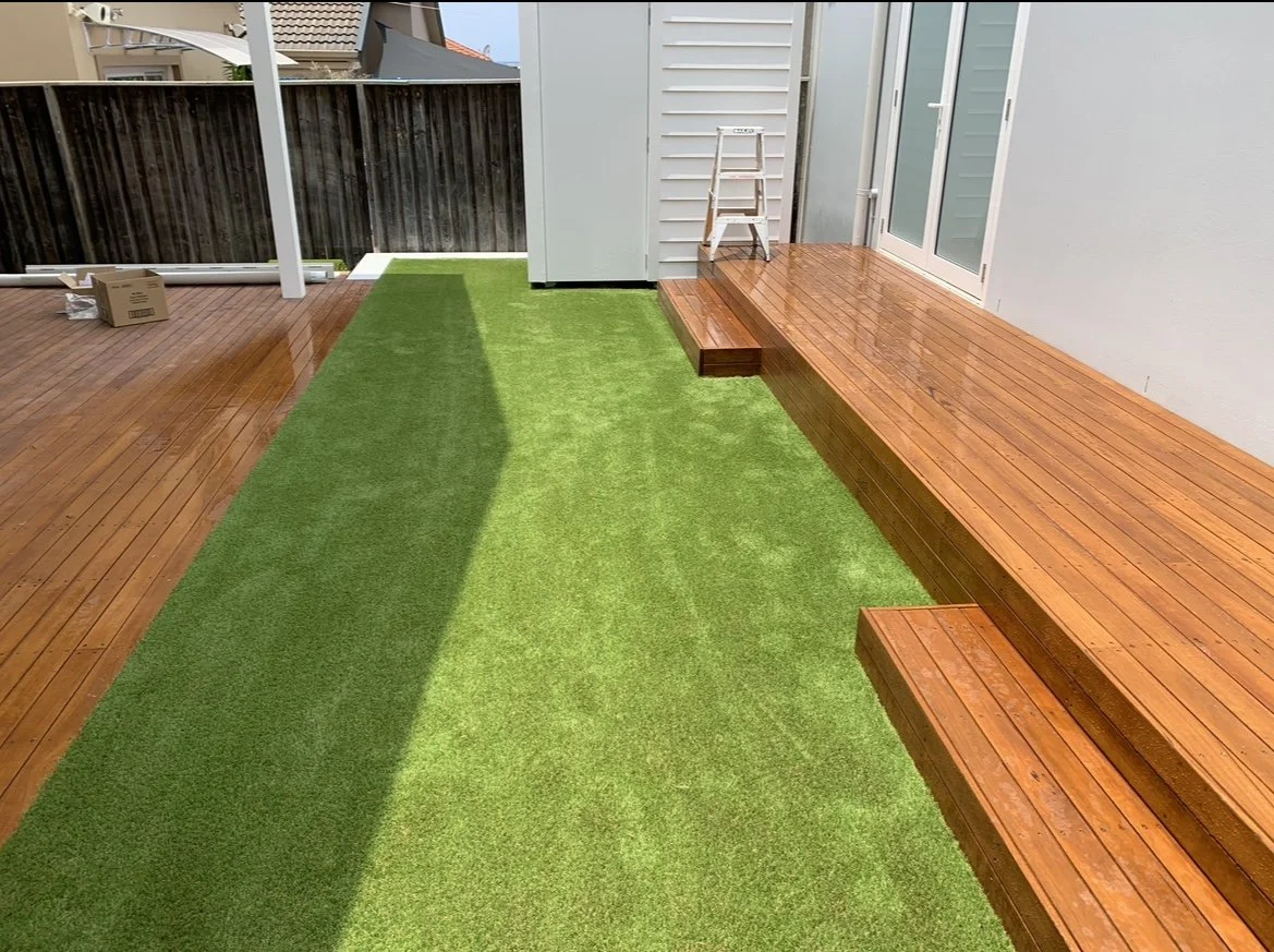 Outdoor deck area with wooden flooring, a small green artificial grass patch, a white ladder, and a sliding glass door.
