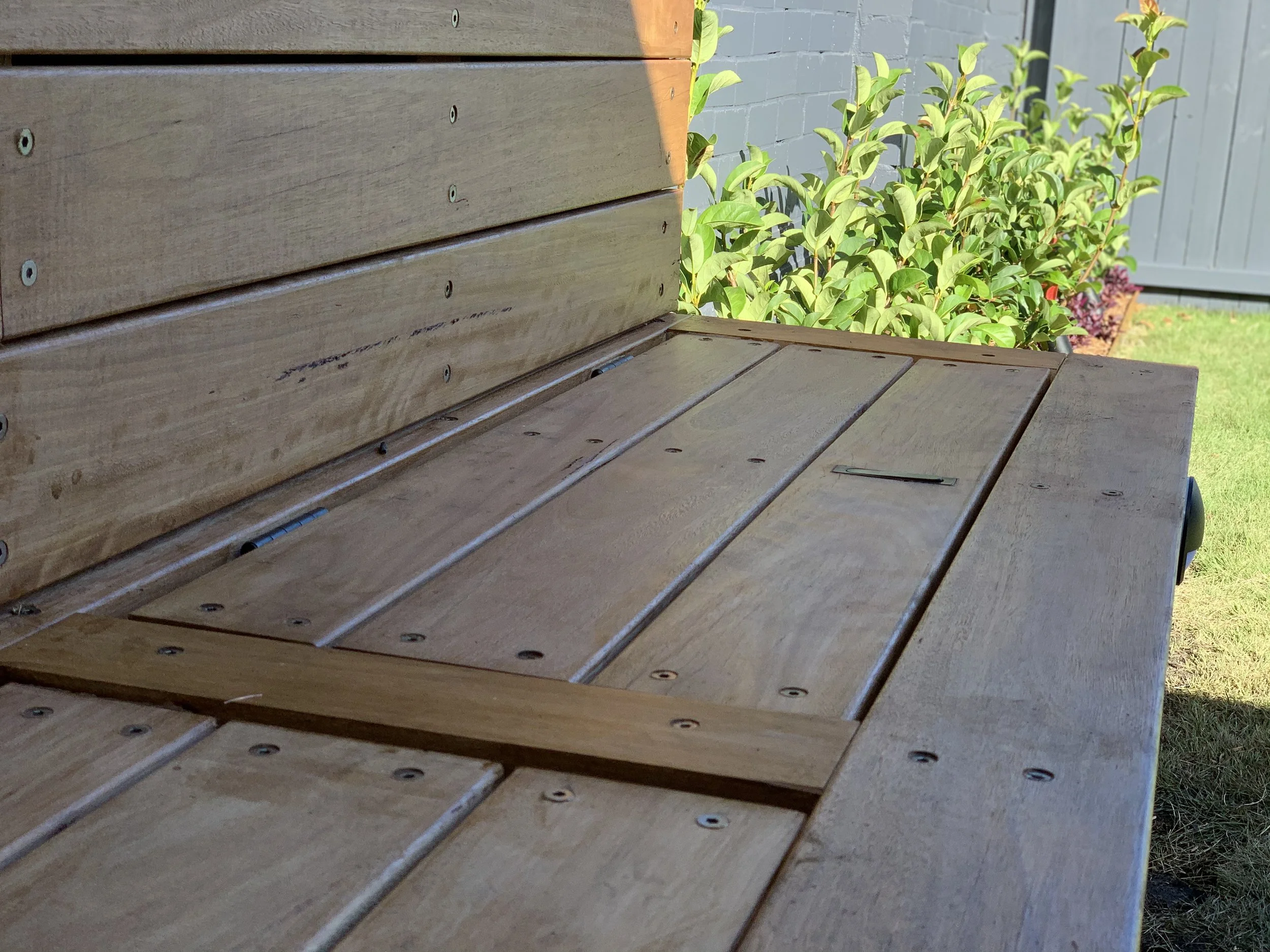 Close-up view of a wooden outdoor bench or table with horizontal wooden planks and visible screws, next to a green leafy shrub and a gray wooden fence.