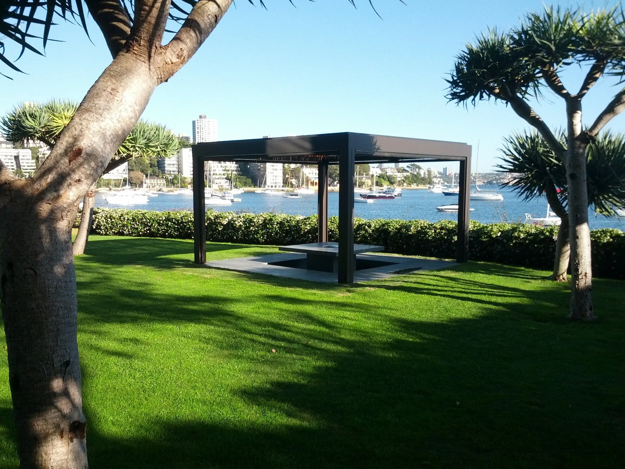 A park overlooking a body of water with boats. There are trees, a grassy area, and a modern black shelter in the park, with tall buildings visible in the distance.